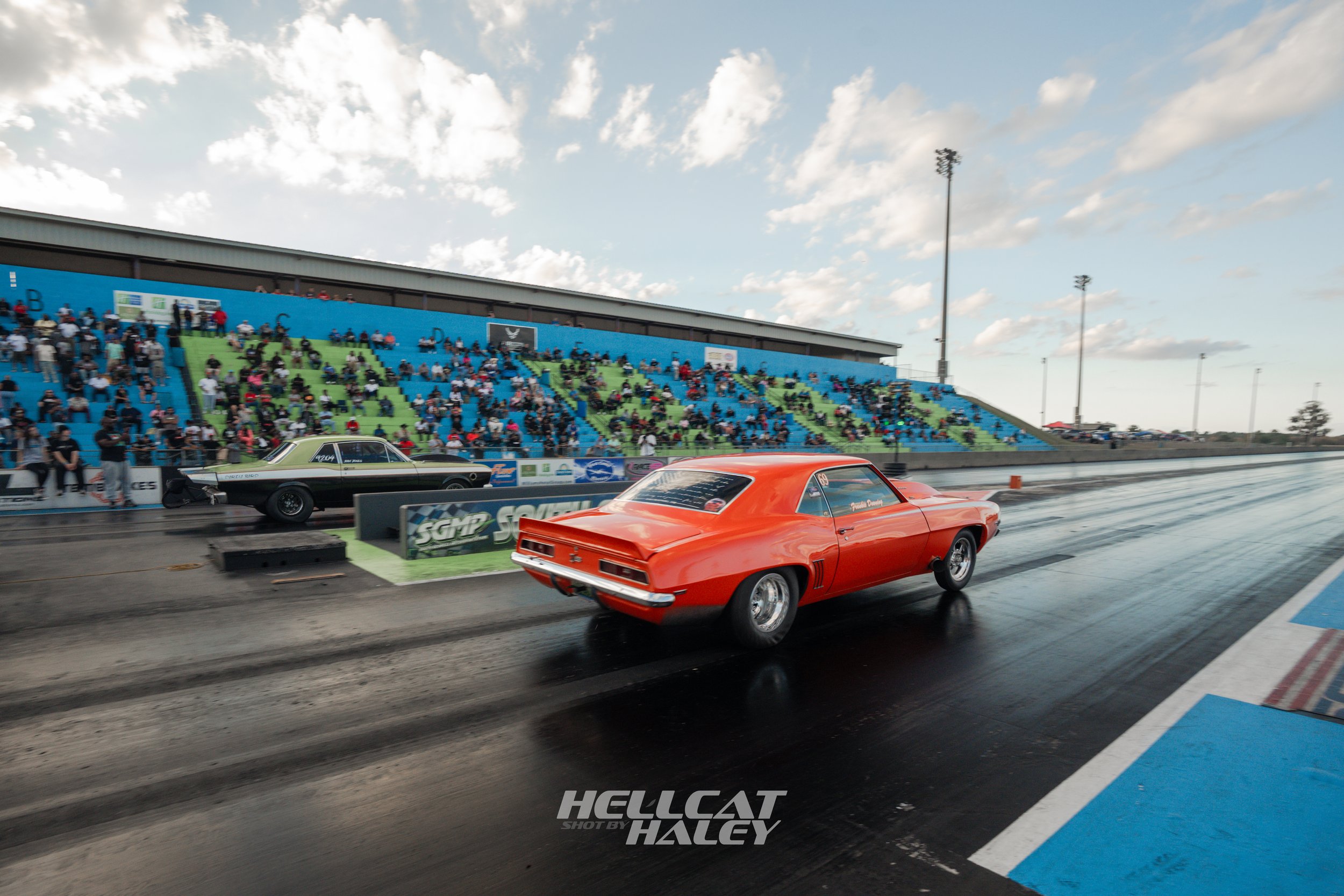 Race cars on a drag strip at a drag racing event with spectators in the stands and a blue sky with clouds overhead.