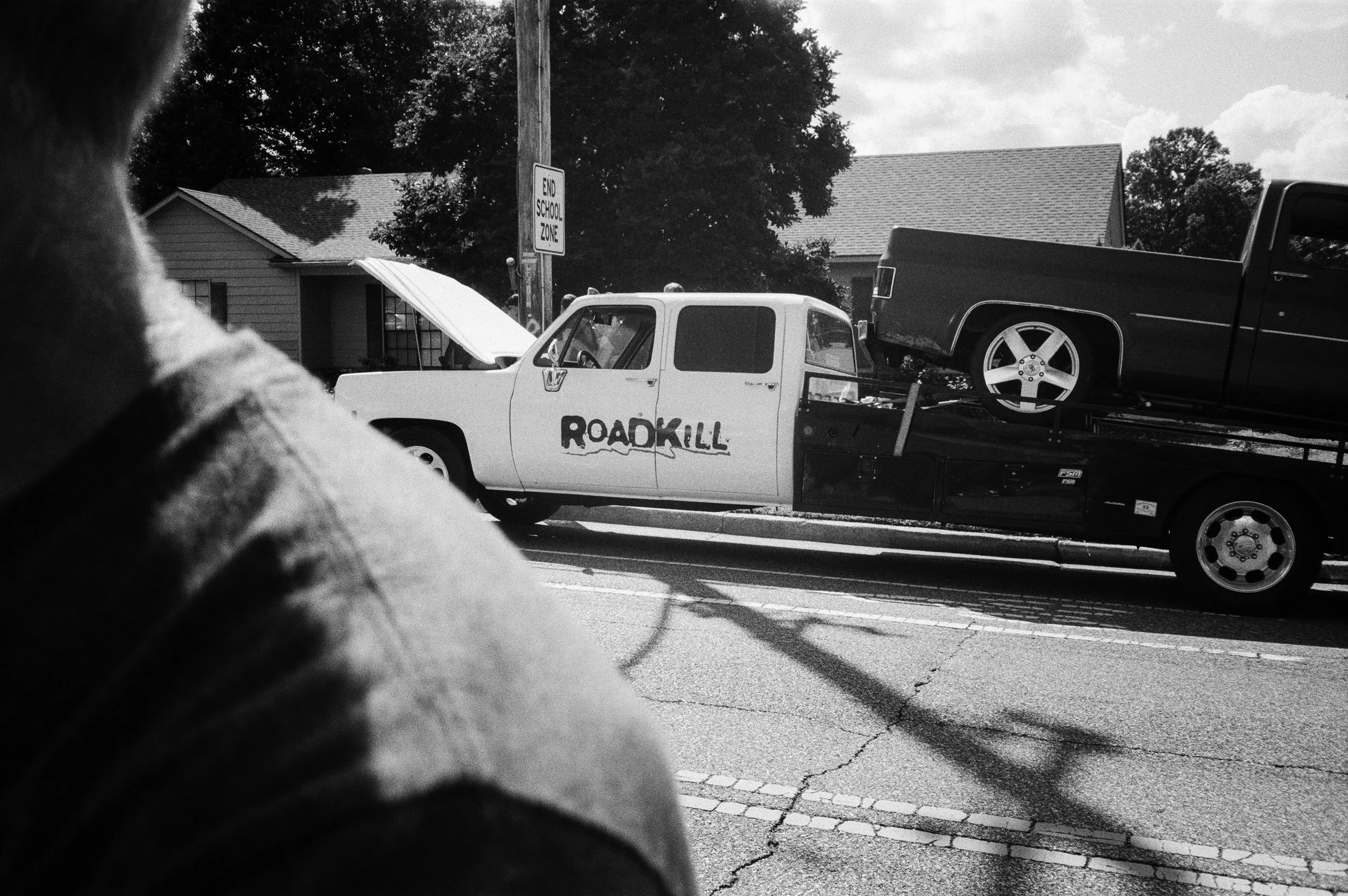 A black and white photo taken from a low angle. In the foreground, part of a person's arm and hand are visible resting on a surface. Behind, on a city street, there is a tow truck with the word 'ROADKILL' written on its door. The truck is carrying a 