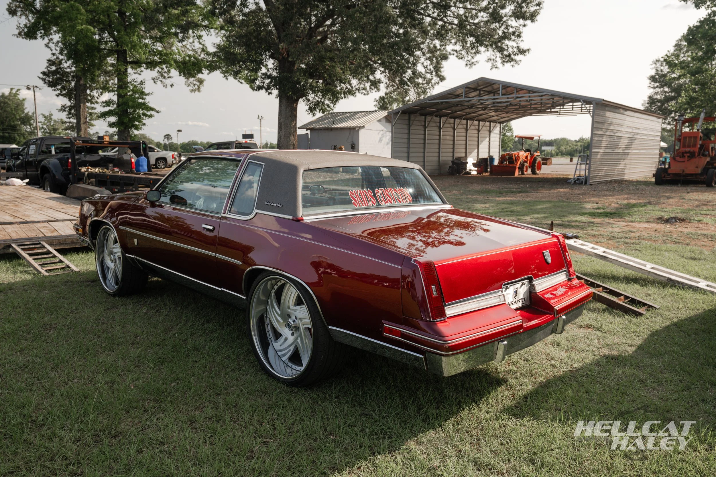 A vintage red car with a gray vinyl roof and large custom wheels parked on grass at an outdoor event with trees, a shed, and a tractor in the background.