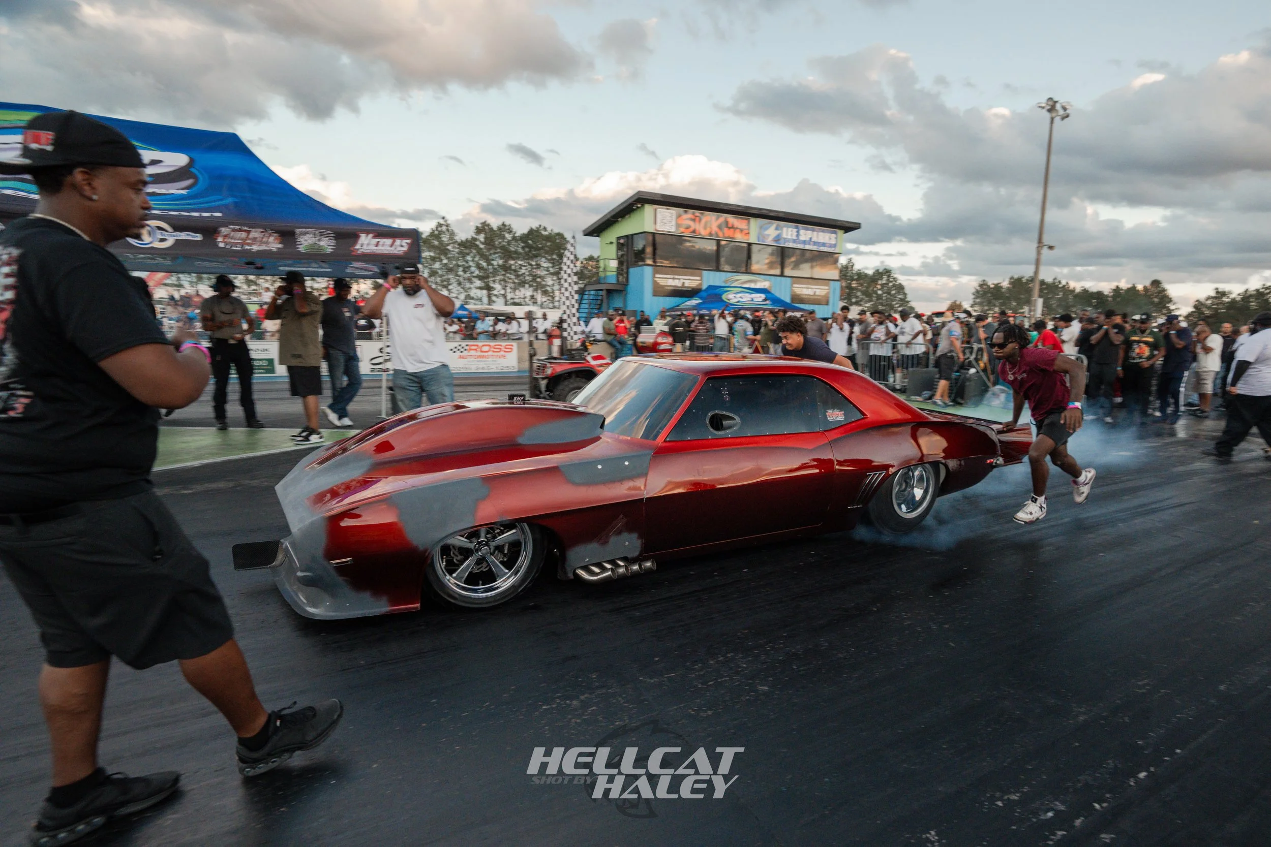 A red race car is on a drag strip with smoke coming from its tires as people watch and take photos in the background. The sky is partly cloudy, and there are tents and a timing tower at the race track.