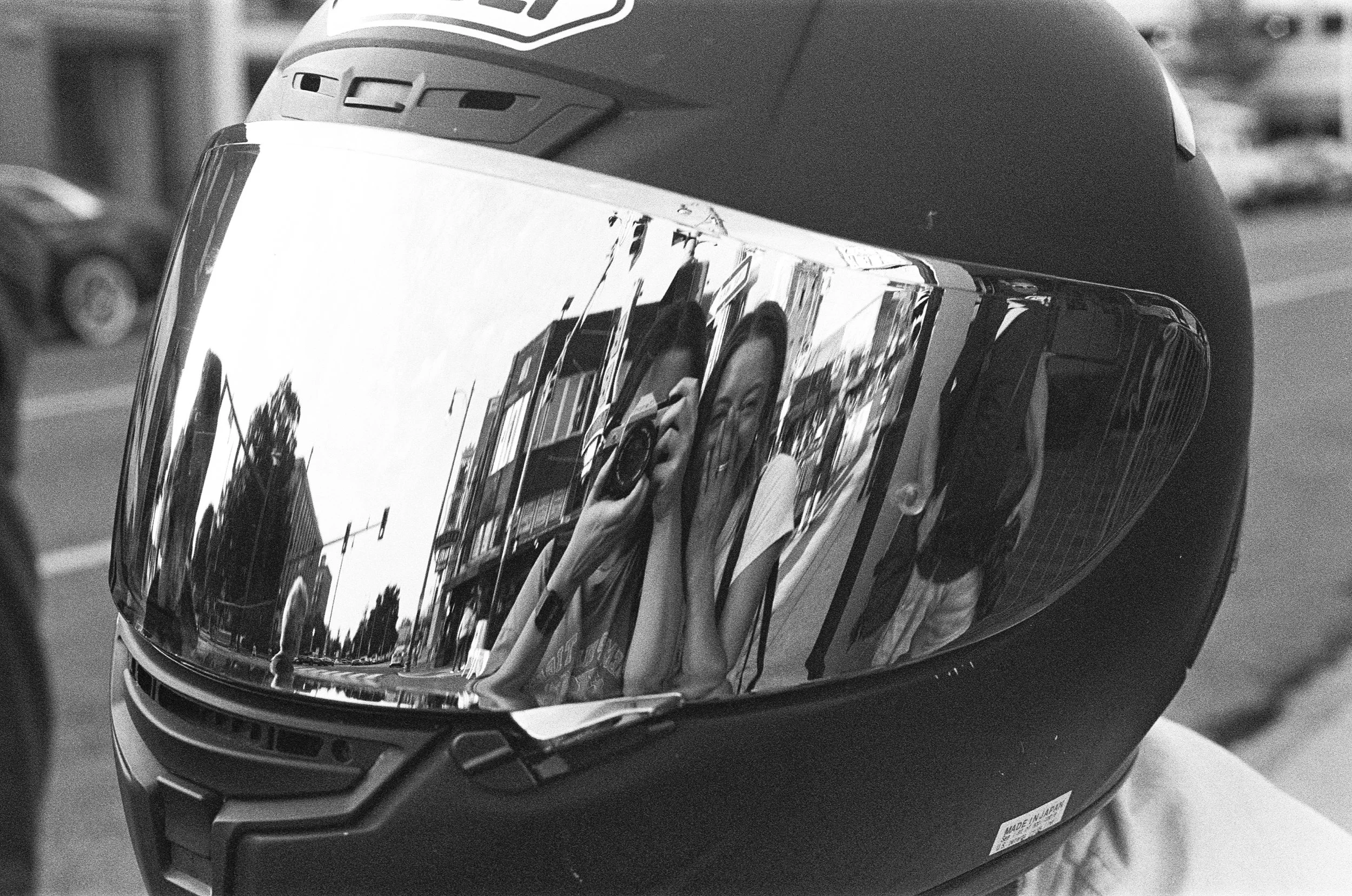 Close-up of a motorcycle helmet with a reflective visor showing a person taking a photo, and a background of a street scene with cars.