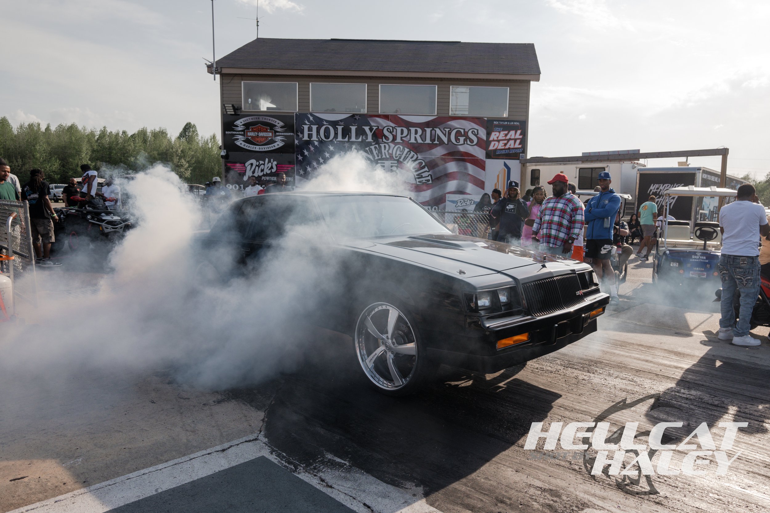 A black car performing a burnout, emitting smoke from the tires at a car event, with a crowd of people and a building with banners in the background.