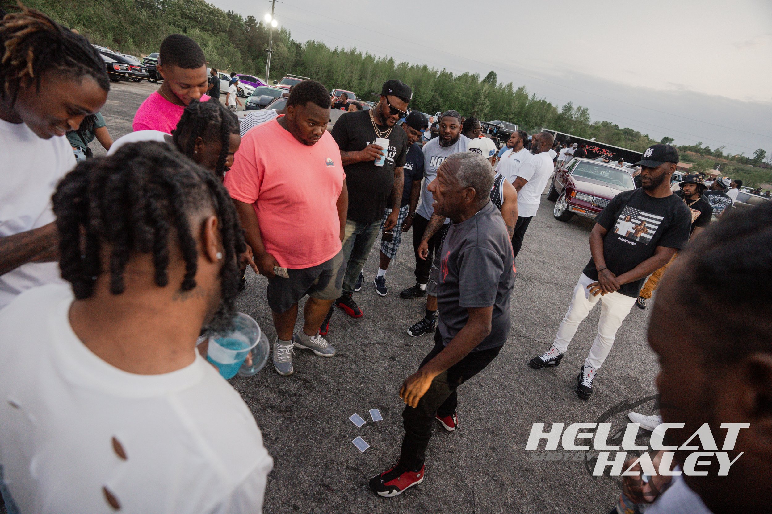 A group of people gathered outdoors in a parking lot, focused on a card game or gambling game on the ground, with several cars parked in the background and green trees beyond.