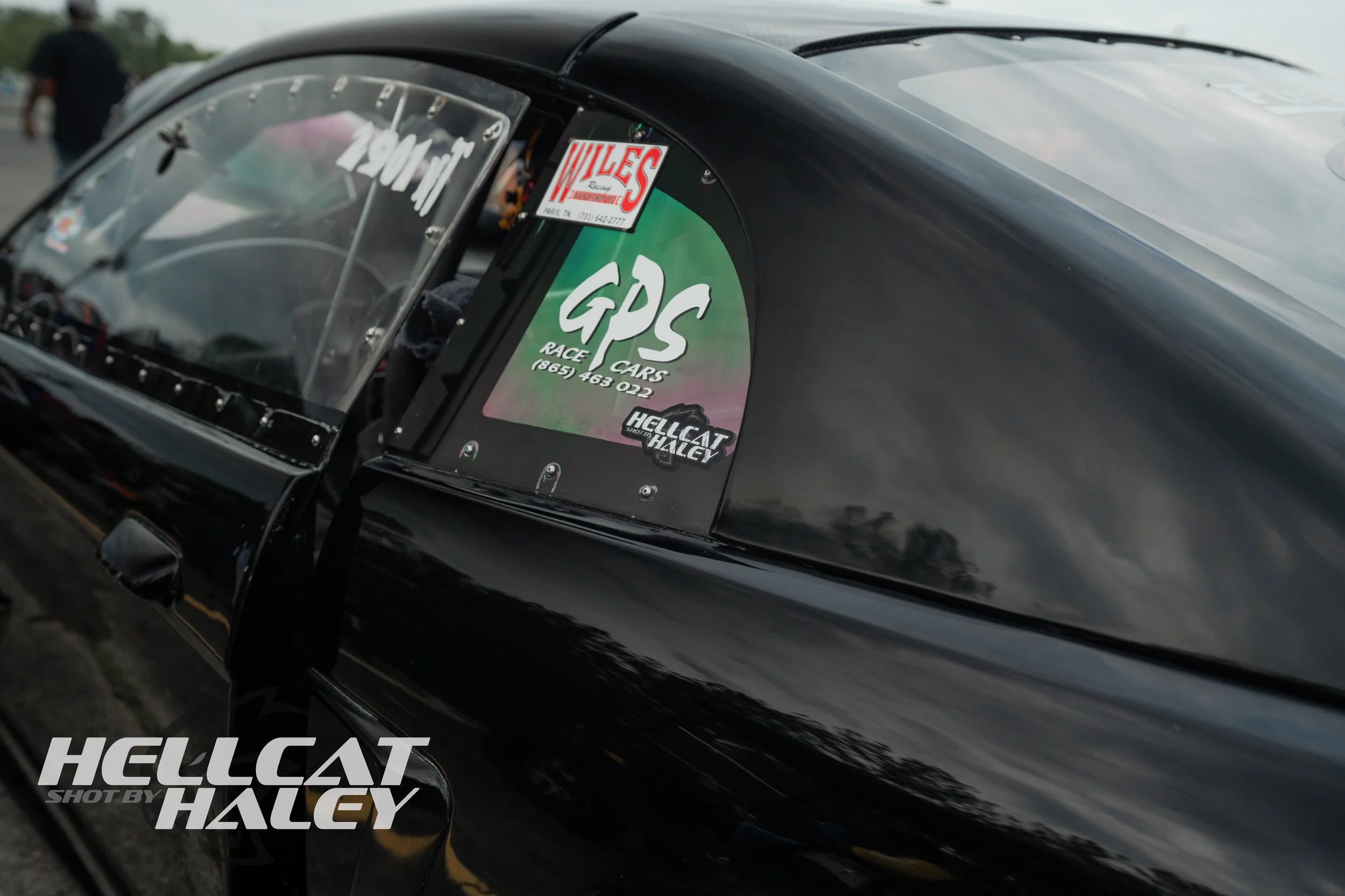 Close-up of a race car's windshield with stickers, including 'GPCS,' 'HelliCAT,' and 'Kyles Racing.' The car is black with reflections on its surface, and a person is visible in the background.