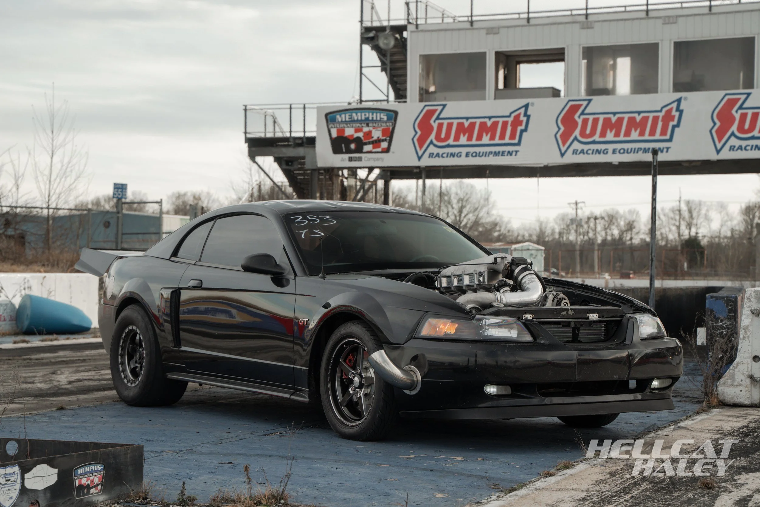 Dismantled black Ford Mustang GT race car with exposed engine parts in a racetrack pit area, with a large sign for Memphis Racing Equipment and Summit Racing Equipment in the background.