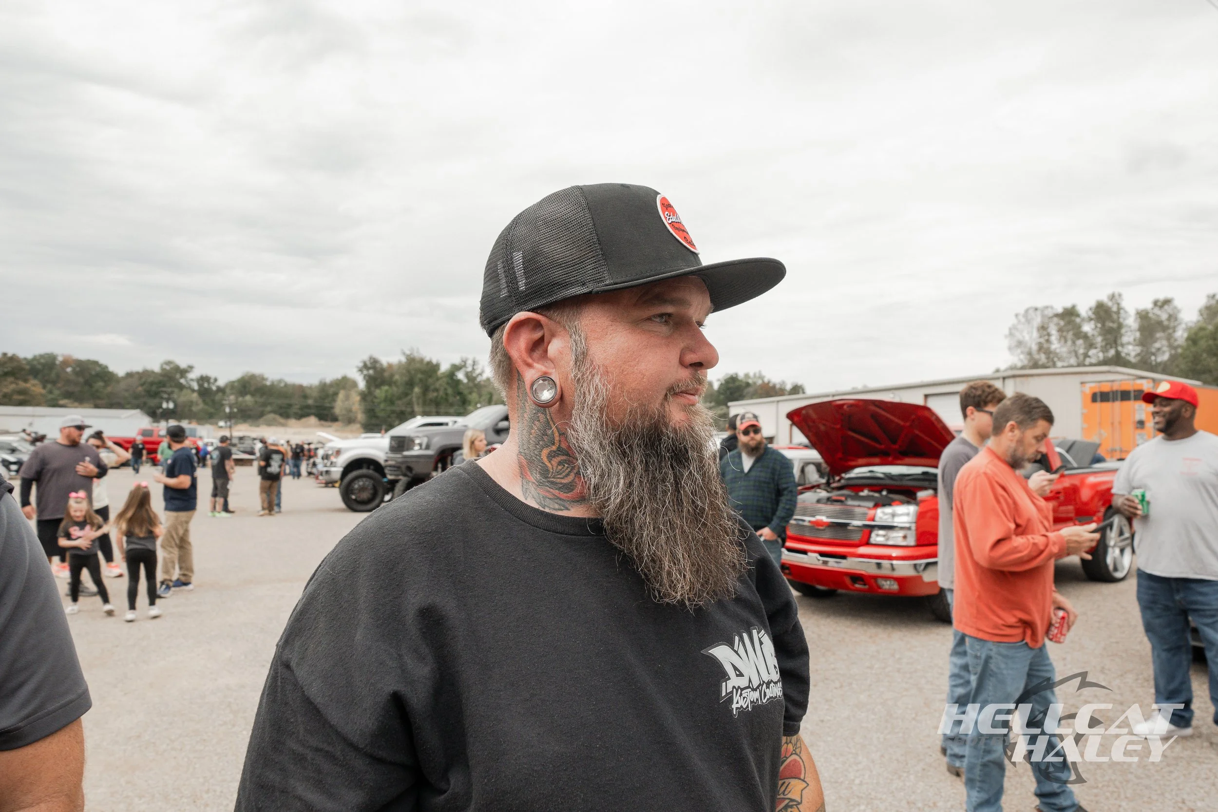 Profile of a man with a gray beard, tattoos on his neck, and large gauge earrings, wearing a black cap and black T-shirt, at an outdoor car event with people, vehicles, and overcast sky in background.