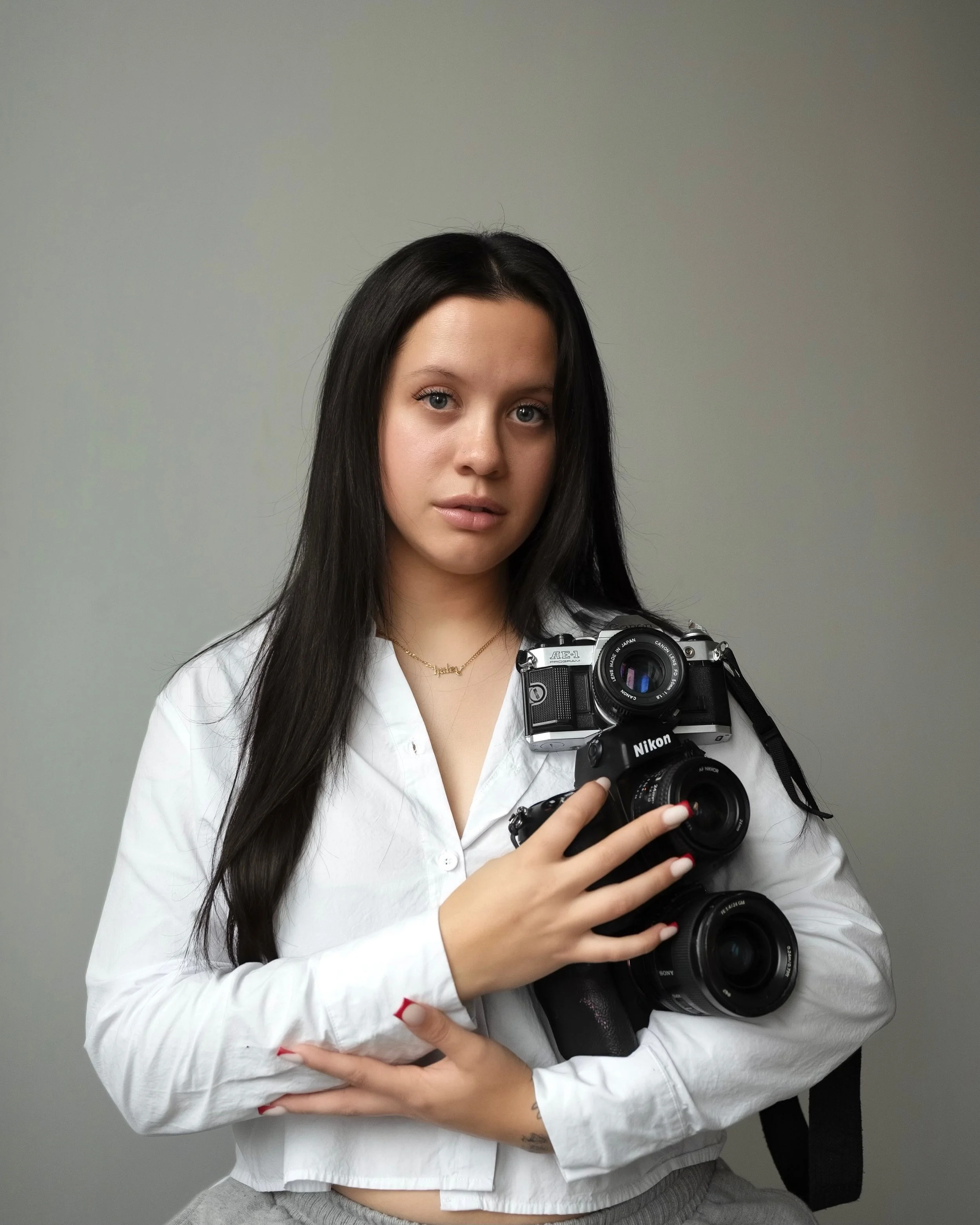 A young woman with long dark hair and light skin taking a selfie with a camera, wearing a white shirt and gray pants, standing against a plain gray background.
