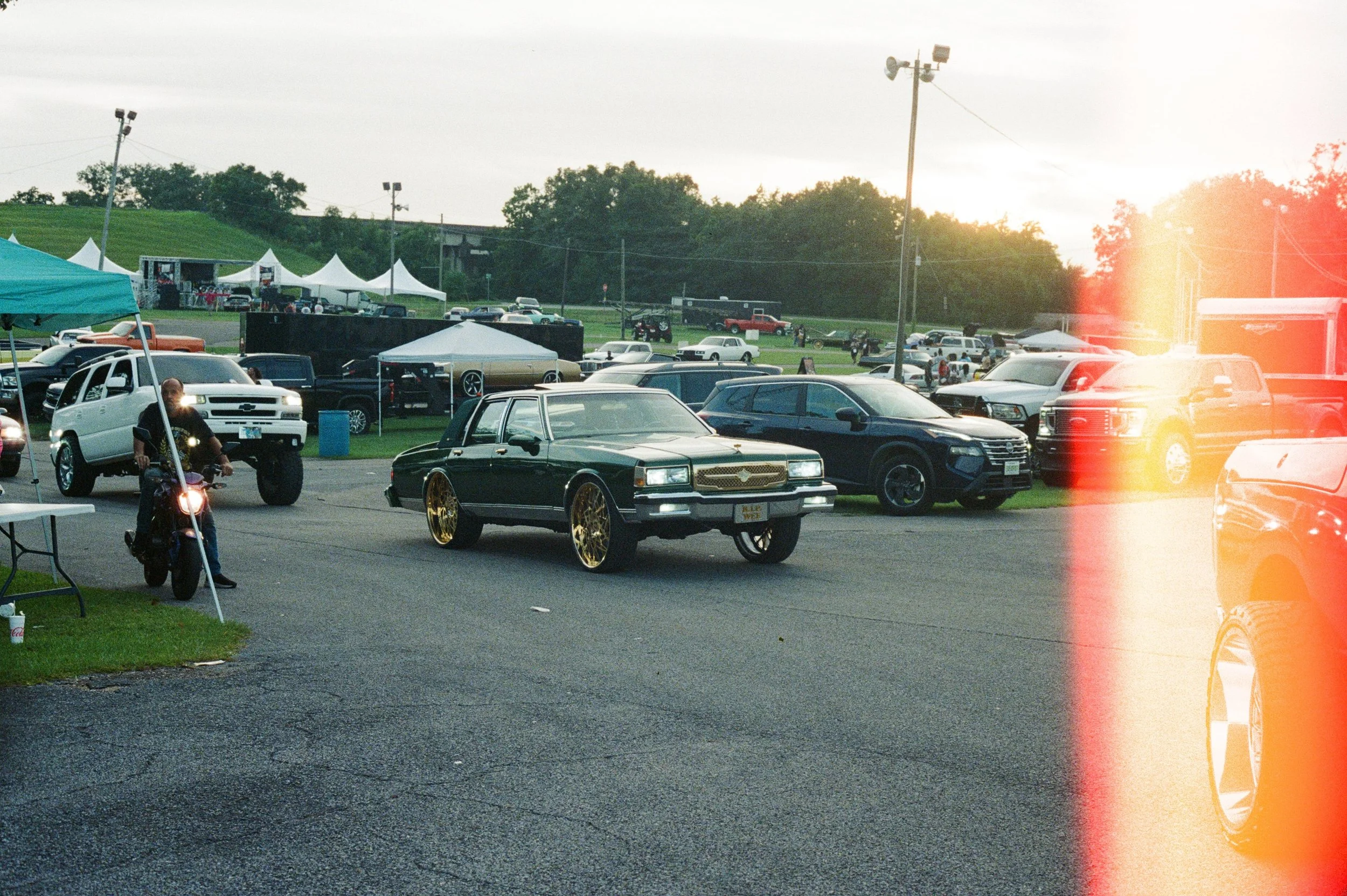 A parking lot at an outdoor event with various cars, tents, and a stage in the background during sunset, with lens flare and a light leak effect on the right side.