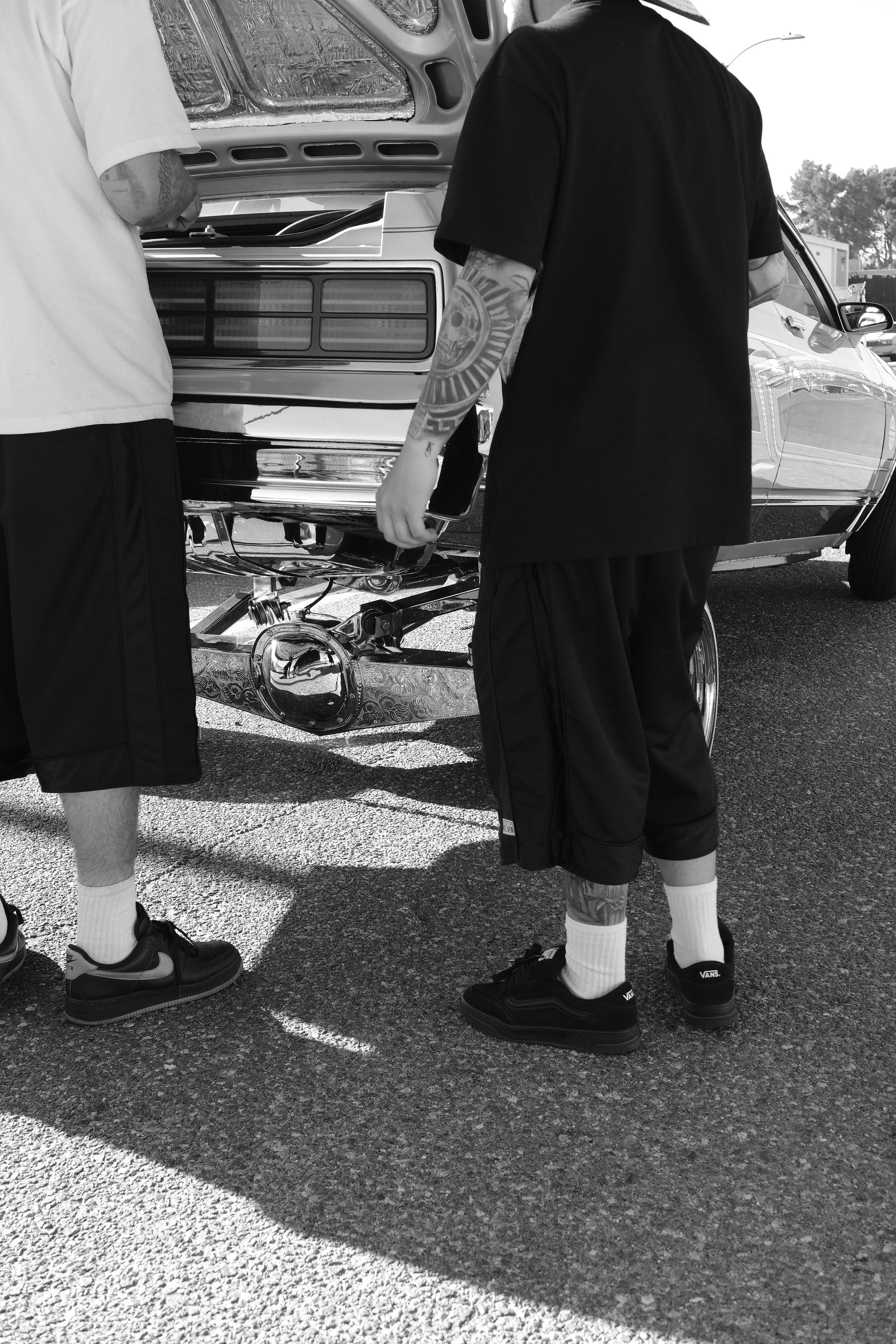 Two people standing next to a car, with the hood open, working on the engine. Only their torsos and legs are visible; one is wearing a white shirt, and the other is wearing a black shirt with tattoos on their arms. They are in a parking lot with othe