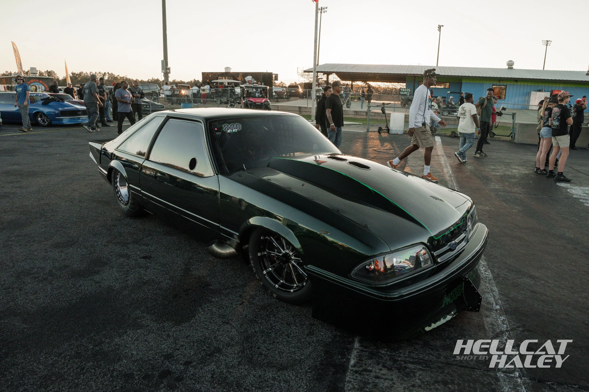 A black vintage Ford Mustang with aftermarket wheels and hood vents parked at a car event, with people walking around and other cars in the background.