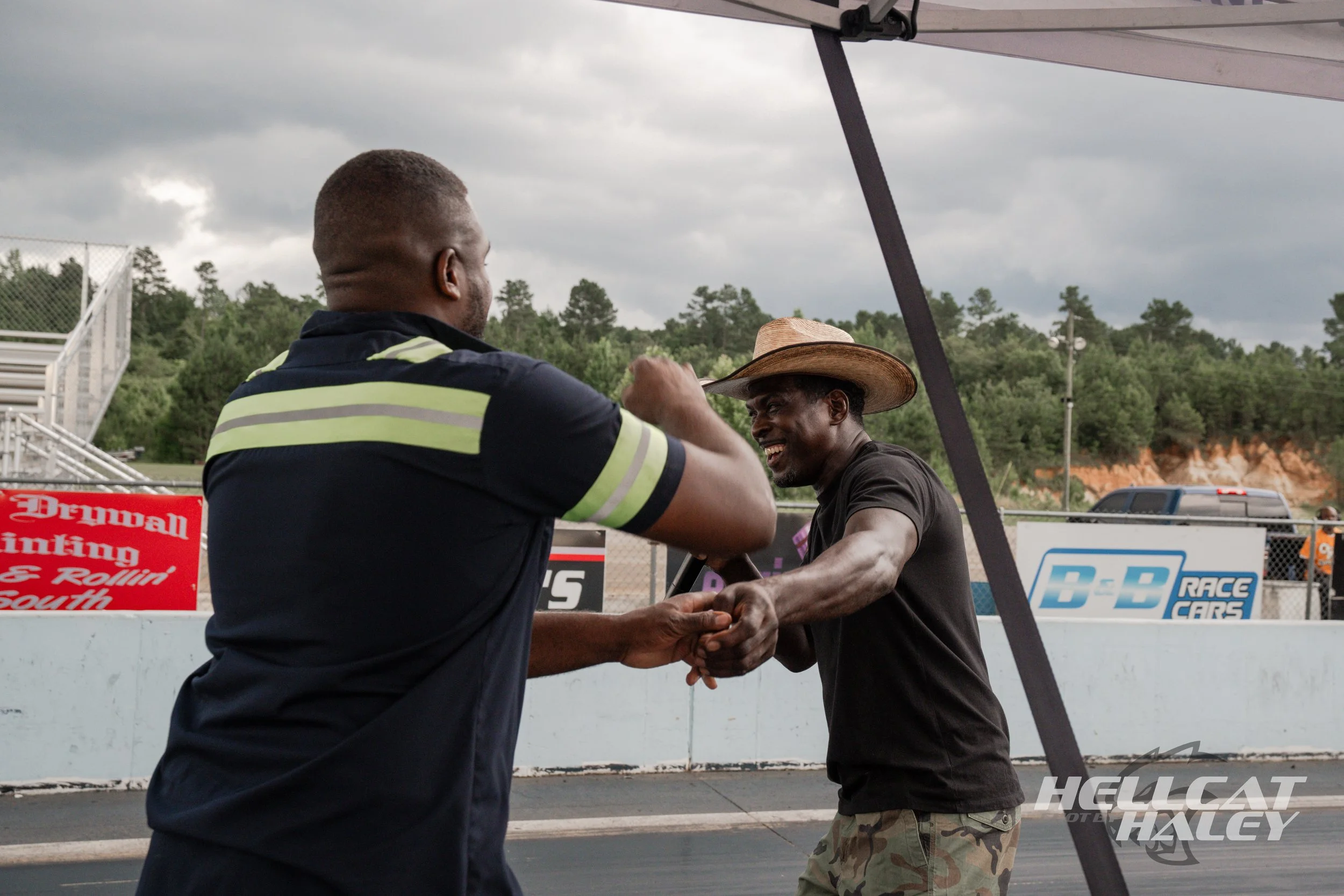 Two men shaking hands at a racetrack under a tent canopy, with one wearing a cowboy hat and camouflage pants, and the other in a sports jersey. Overcast sky and trees in the background.