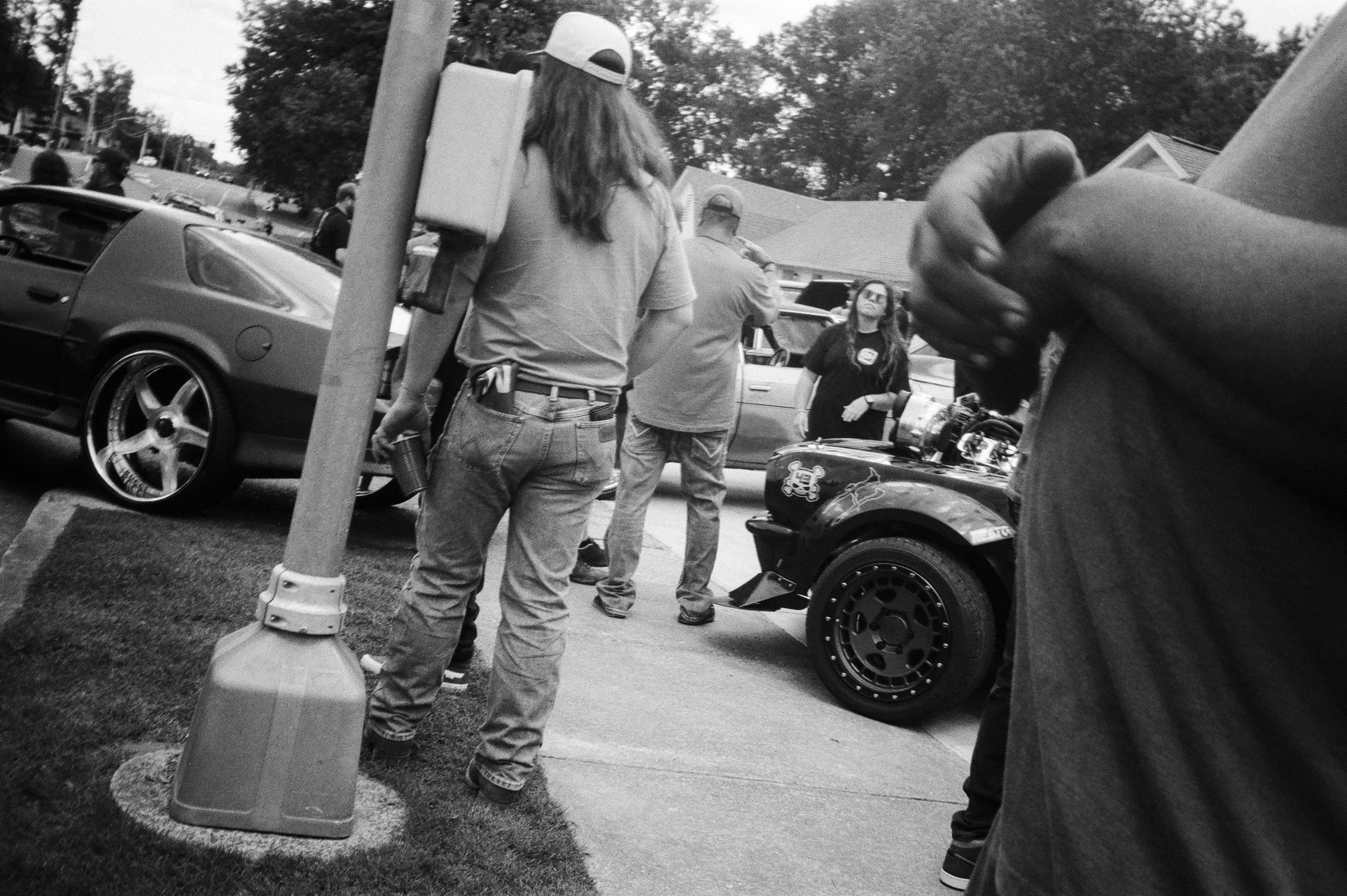 People gathered outdoors around a parked sports car with custom wheels. Some are looking at the car, while others stand nearby. There's a woman with long hair wearing glasses and a T-shirt. The scene appears to be at a casual outdoor event or car sho