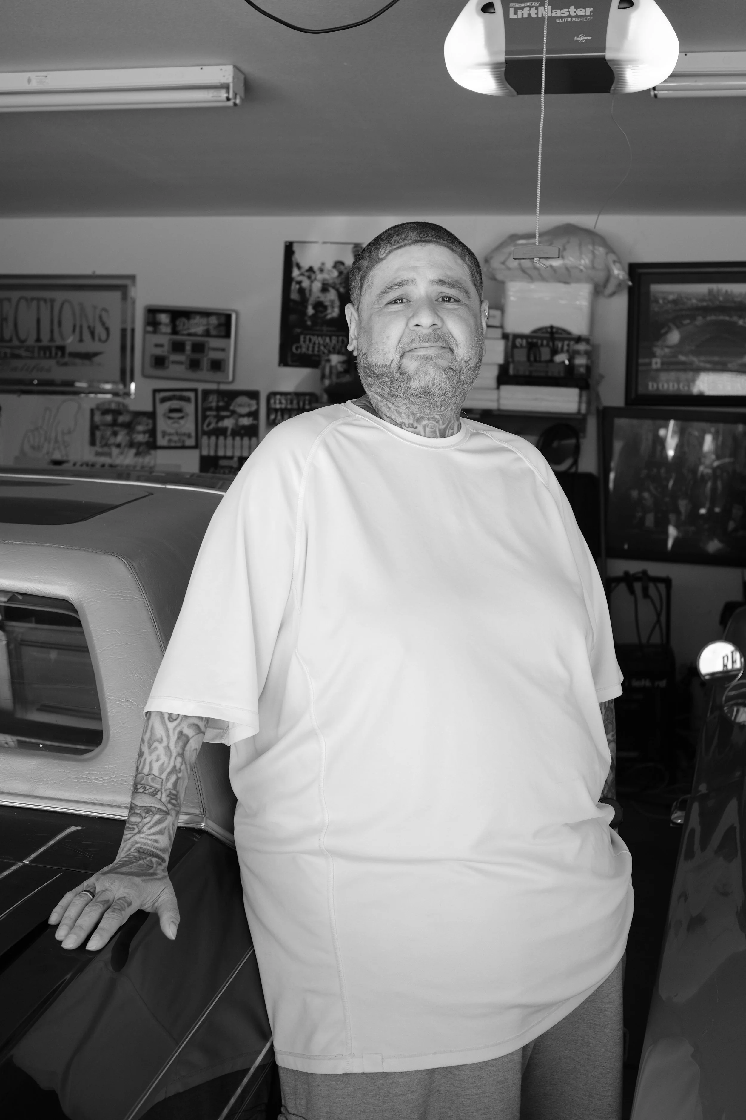 A man with tattoos on his left arm standing indoors with a garage door opener above him, various posters and shelves with books and items in the background, and a television on the right side.