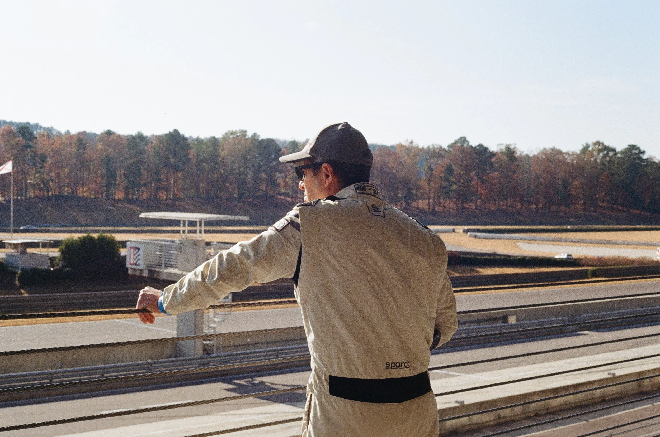 A man in racing gear and sunglasses appears to be on a balcony overlooking a racetrack with trees in the background.