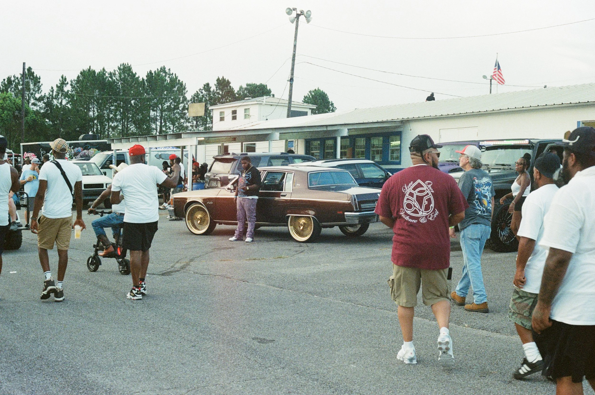 A street scene with a crowd of people, vintage cars, and a building with an American flag in the background.