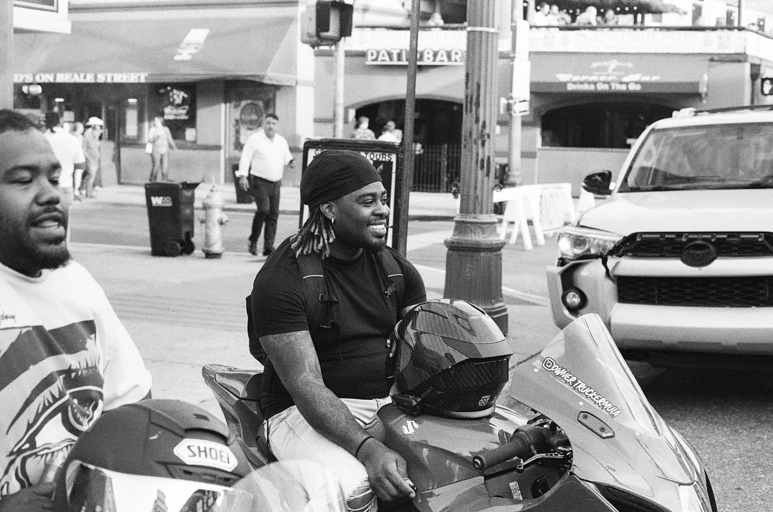 Two men sitting on their motorcycles on a city street, with pedestrians and storefronts in the background. One man is smiling and the other is looking off to the side.