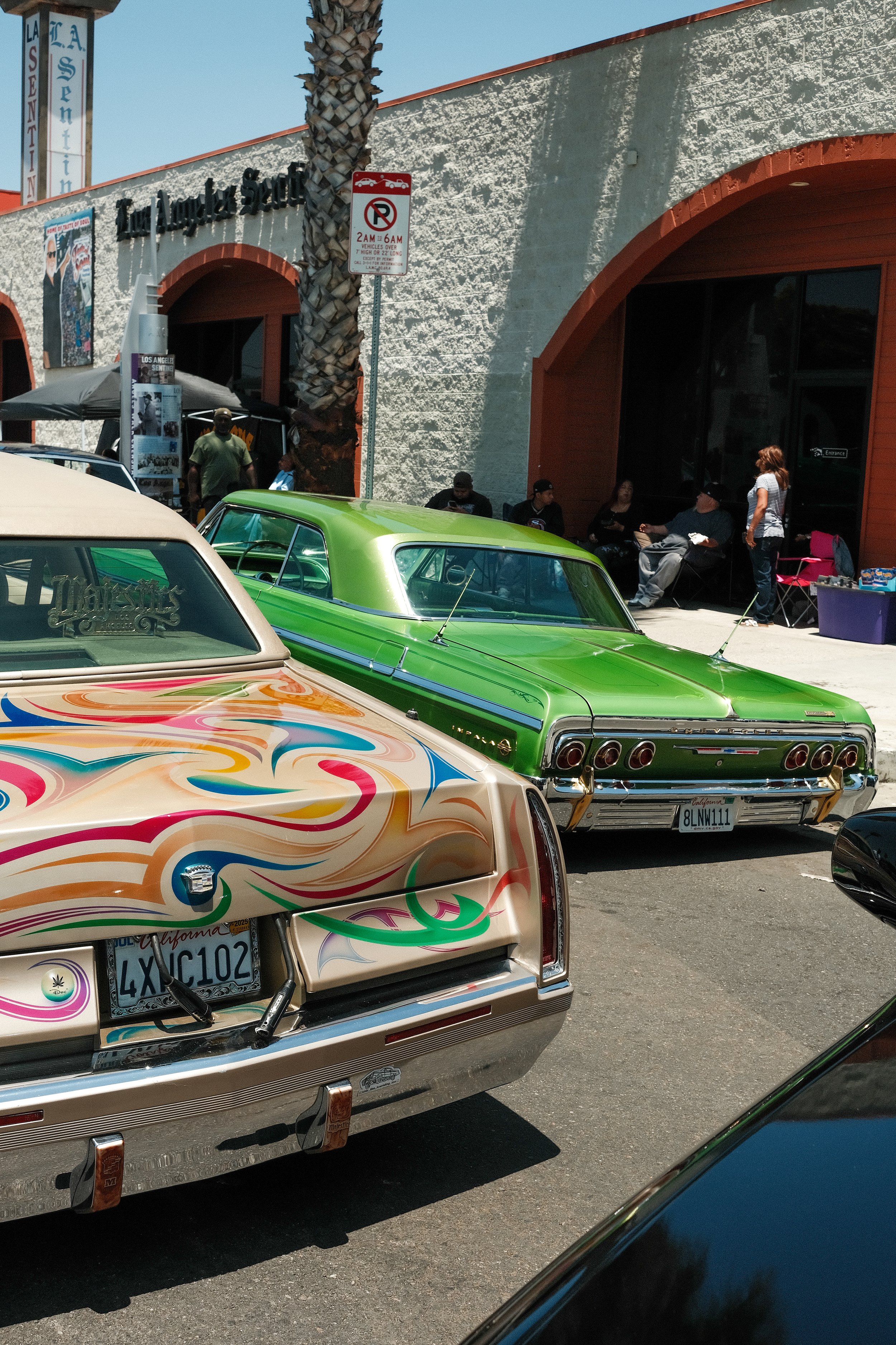 Parking lot with two vintage cars, one beige with colorful flames on the trunk and license plate 4XIC102, and a bright green car behind it. People are sitting and standing near the entrance of a building with a sign that reads "Los Angeles.”