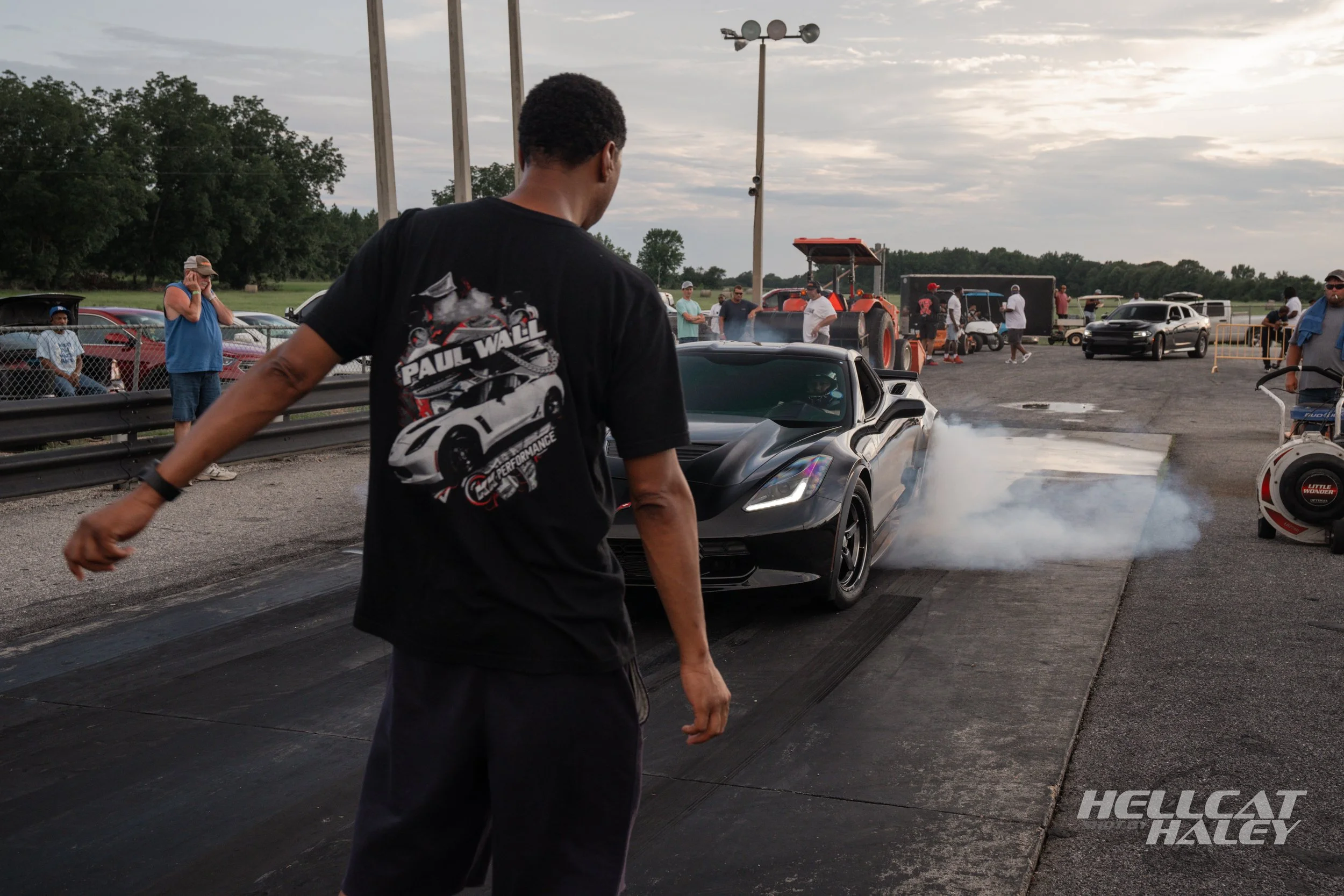 A man wearing a black T-shirt with a car graphic and the name 'Paul Wall' standing behind a black sports car on a drag strip with smoke coming from the tires, spectators watching in the background, and racing equipment around, at a racetrack during l