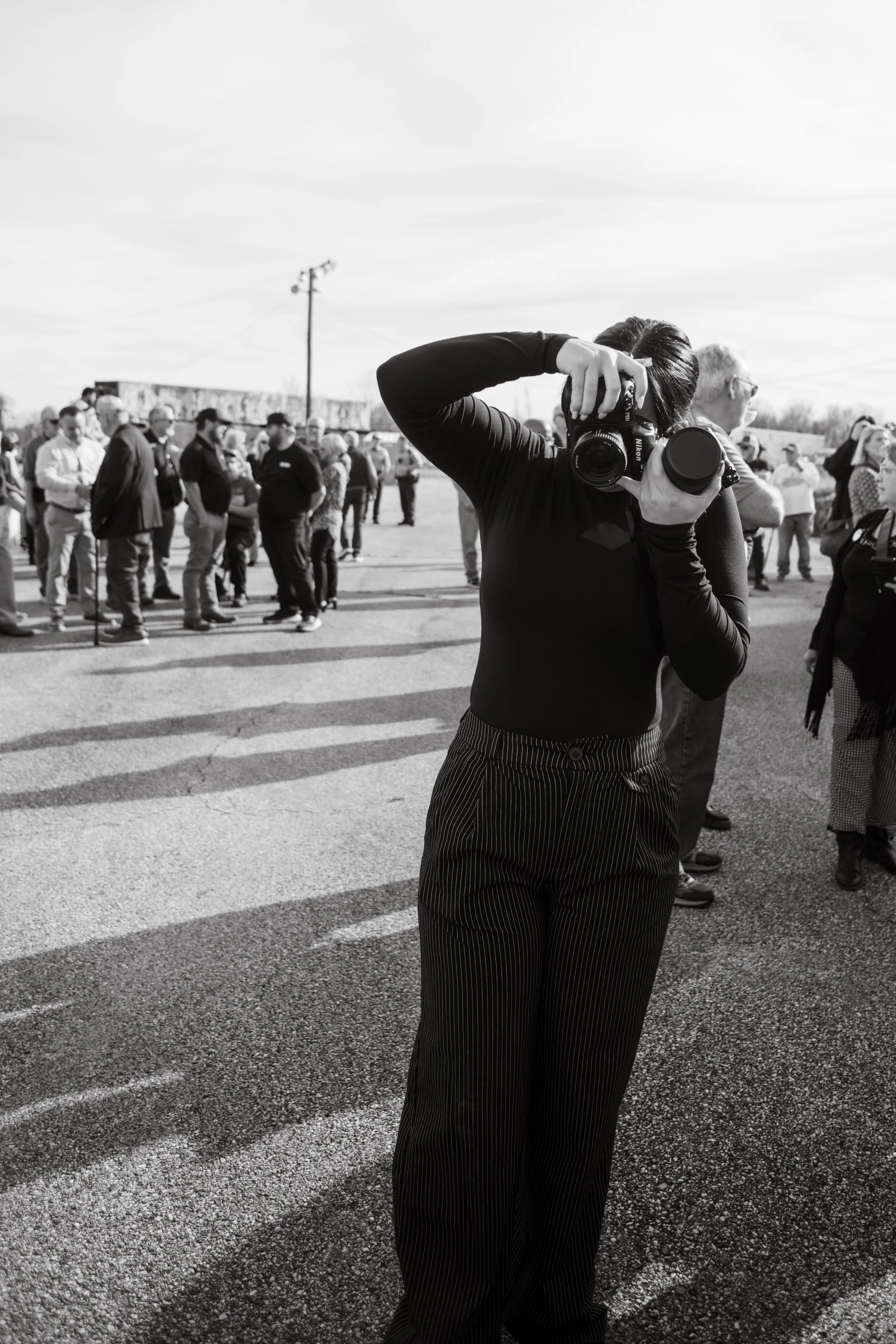 A woman taking a photo with a DSLR camera in a crowd outdoors, with people standing in line and an open sky in the background.