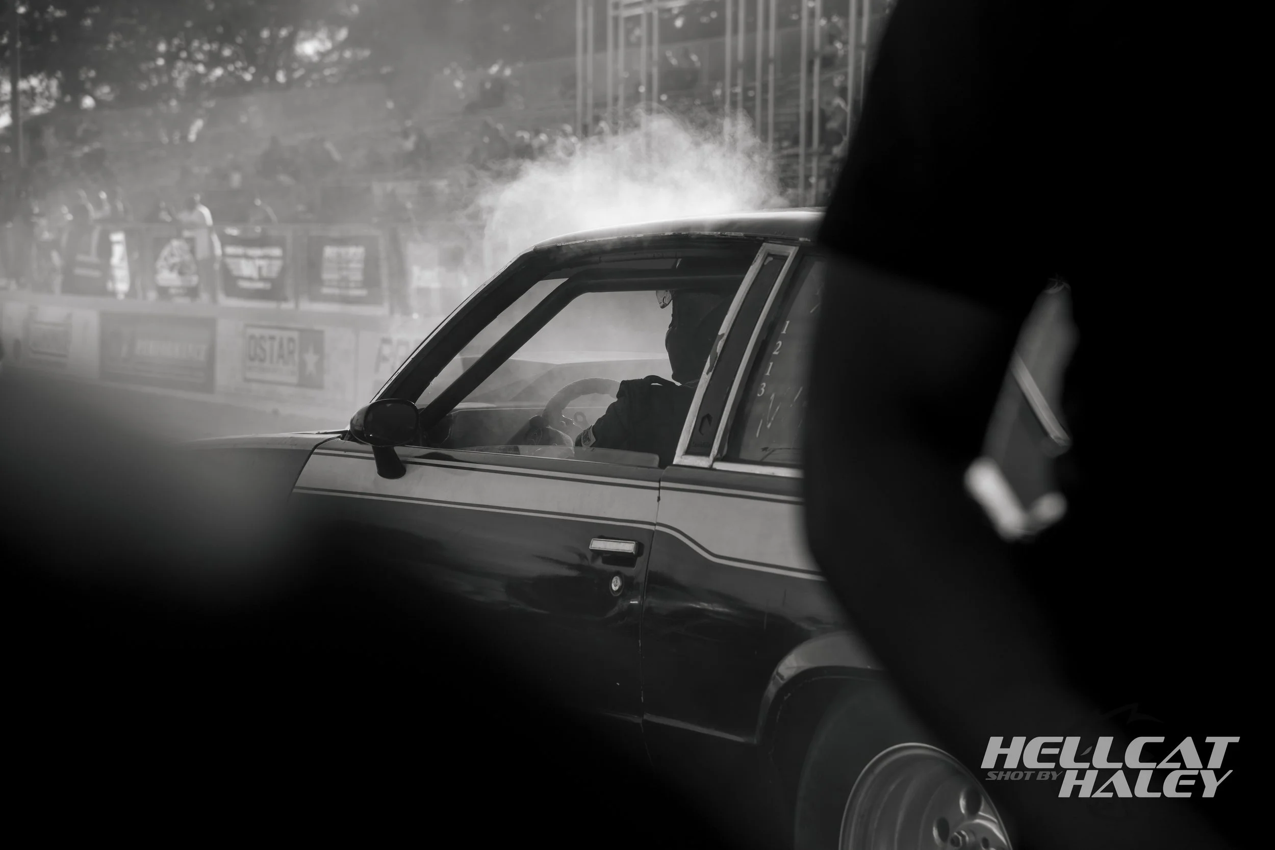 A black-and-white photo captures a race car and driver in the starting position, with a cloud of smoke or vapor near the windshield, possibly from the engine or tires. The background shows a blurred racetrack with advertisements and spectators,