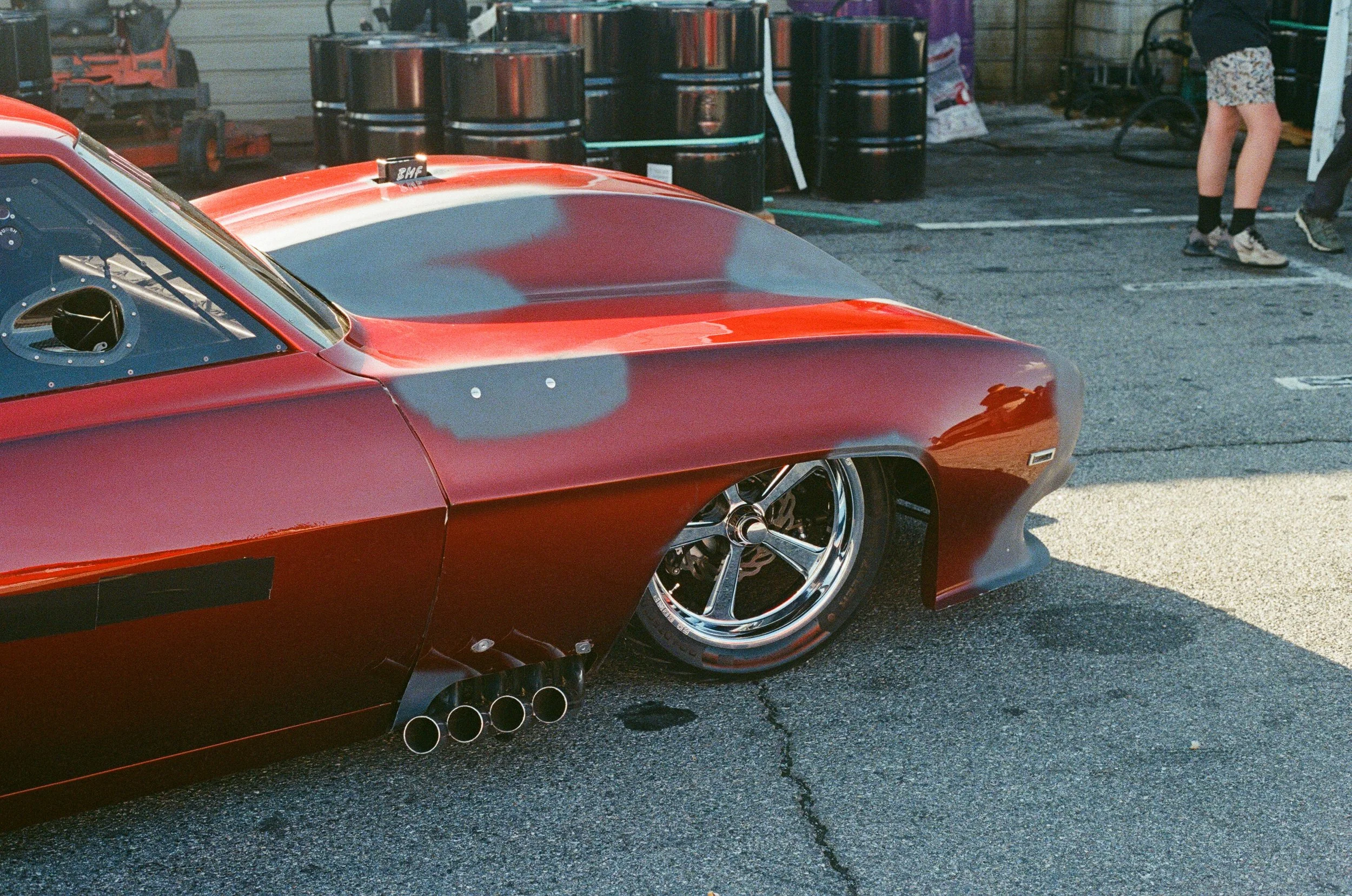 Close-up of a red vintage race car with chrome wheels and exhaust pipes, at a pit stop area with barrels and people in the background.