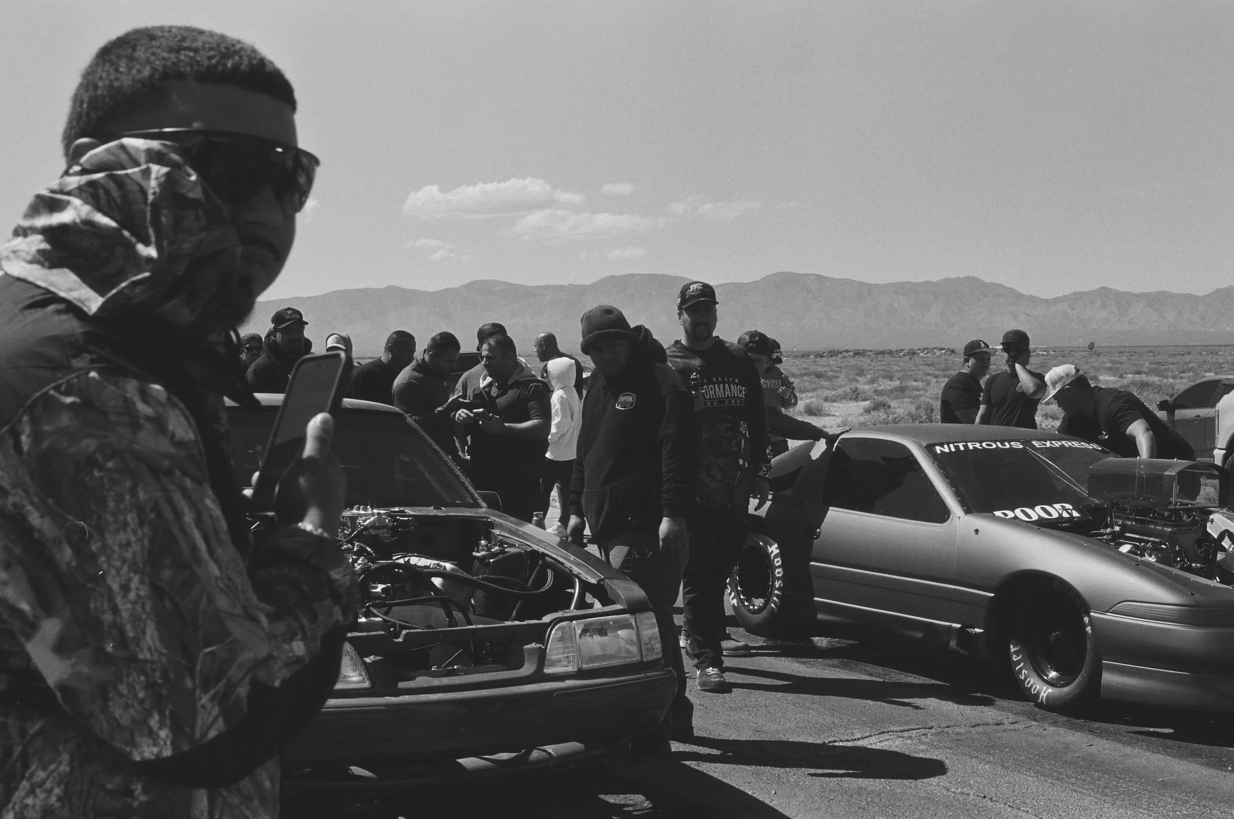 A group of people gathered around cars in a desert landscape with mountains in the background.