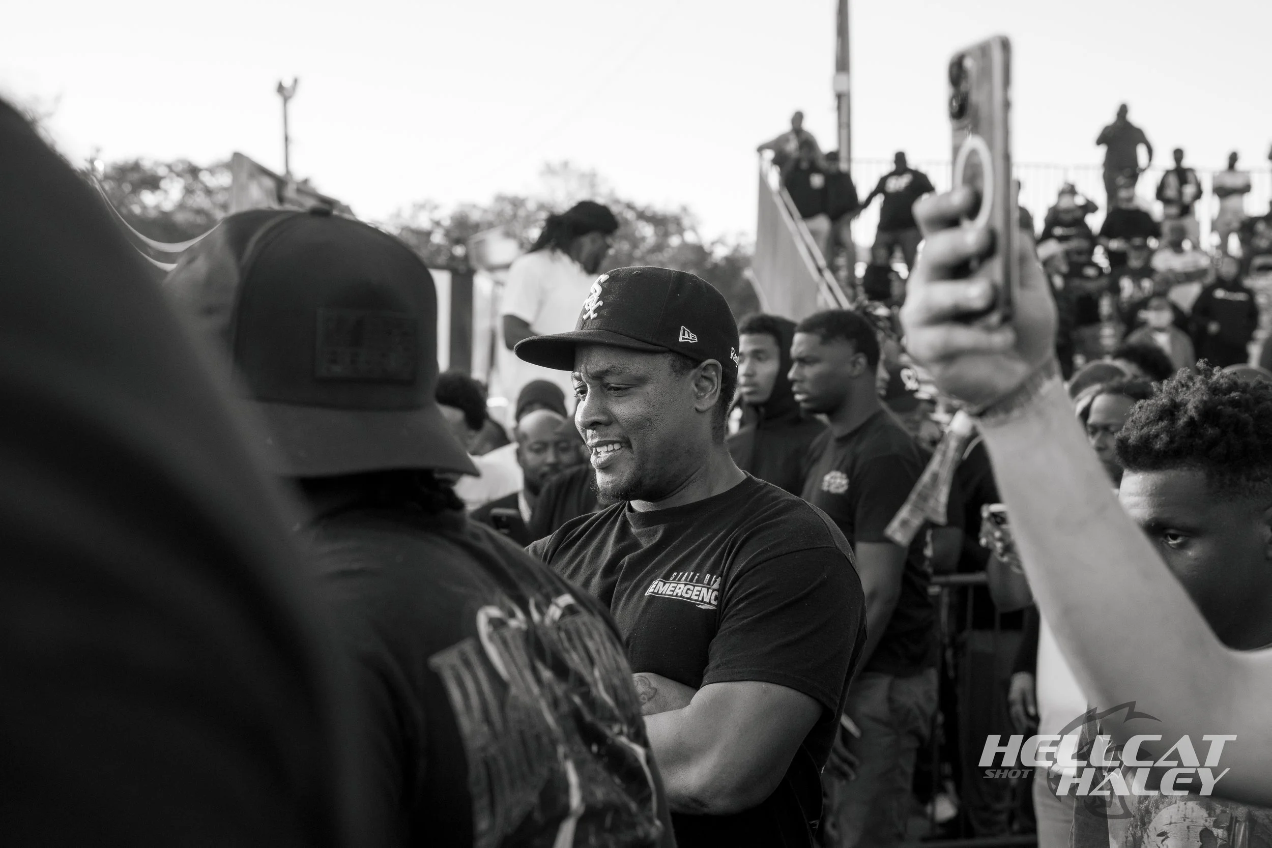 A black and white photograph of a group of people at an outdoor event, with some individuals wearing baseball caps and shirts with logos. A man in the center is smiling, and others around him are looking in different directions. The scene includes a 