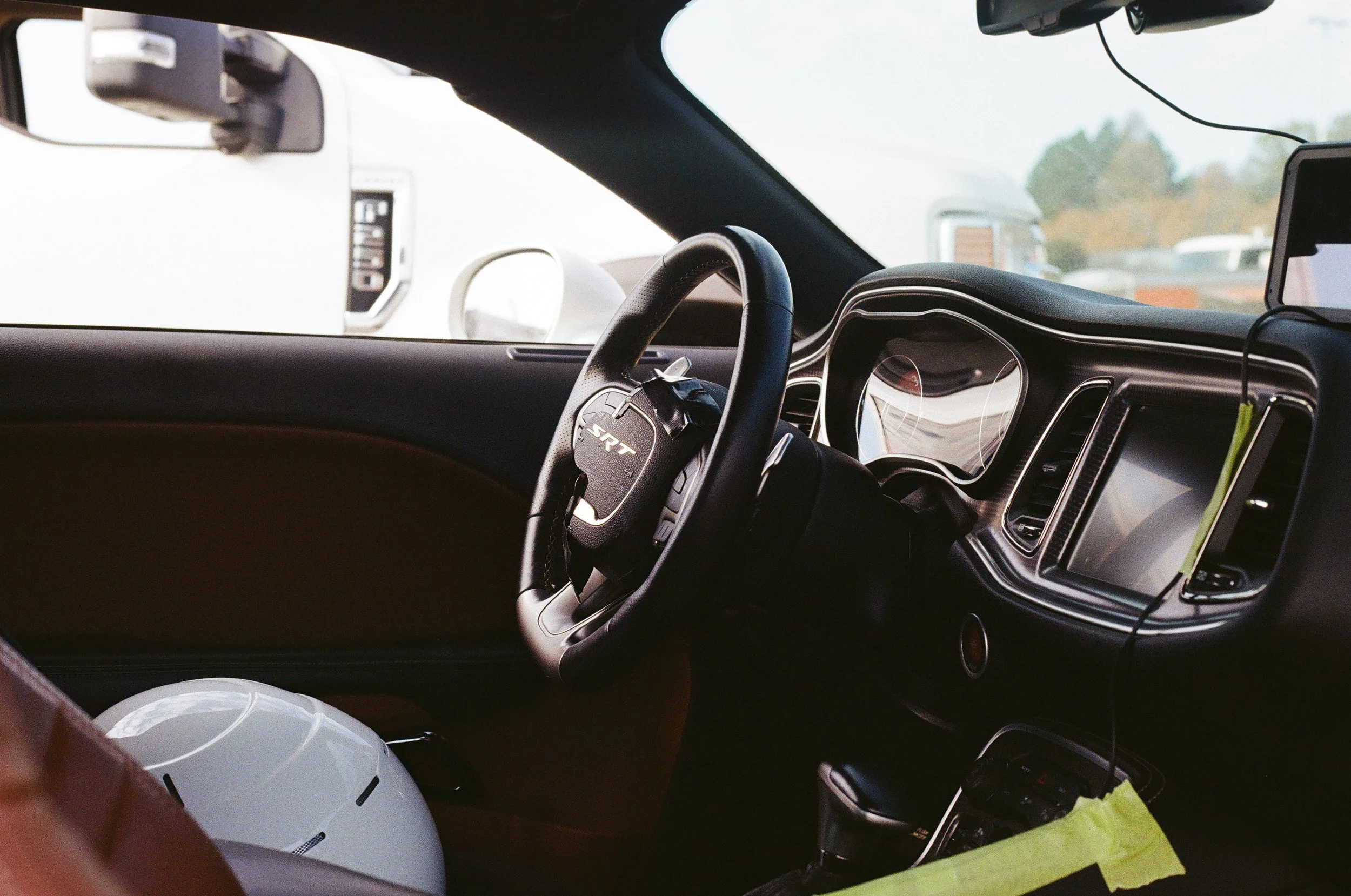 The interior of a car showing the steering wheel, dashboard, and part of the front passenger seat. Outside views through the windshield include other vehicles and trees.