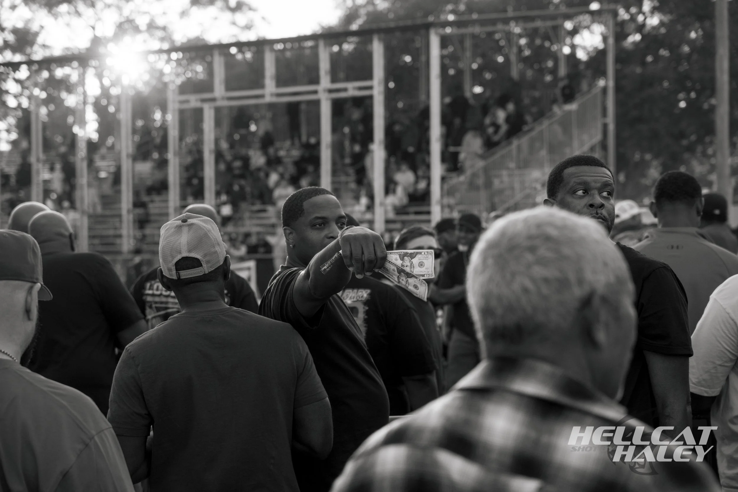 A crowd of people at an outdoor event, some seated on bleachers, with one man in the foreground holding a twenty-dollar bill. The background shows a metal bleacher structure and trees, with sunlight peeking through.