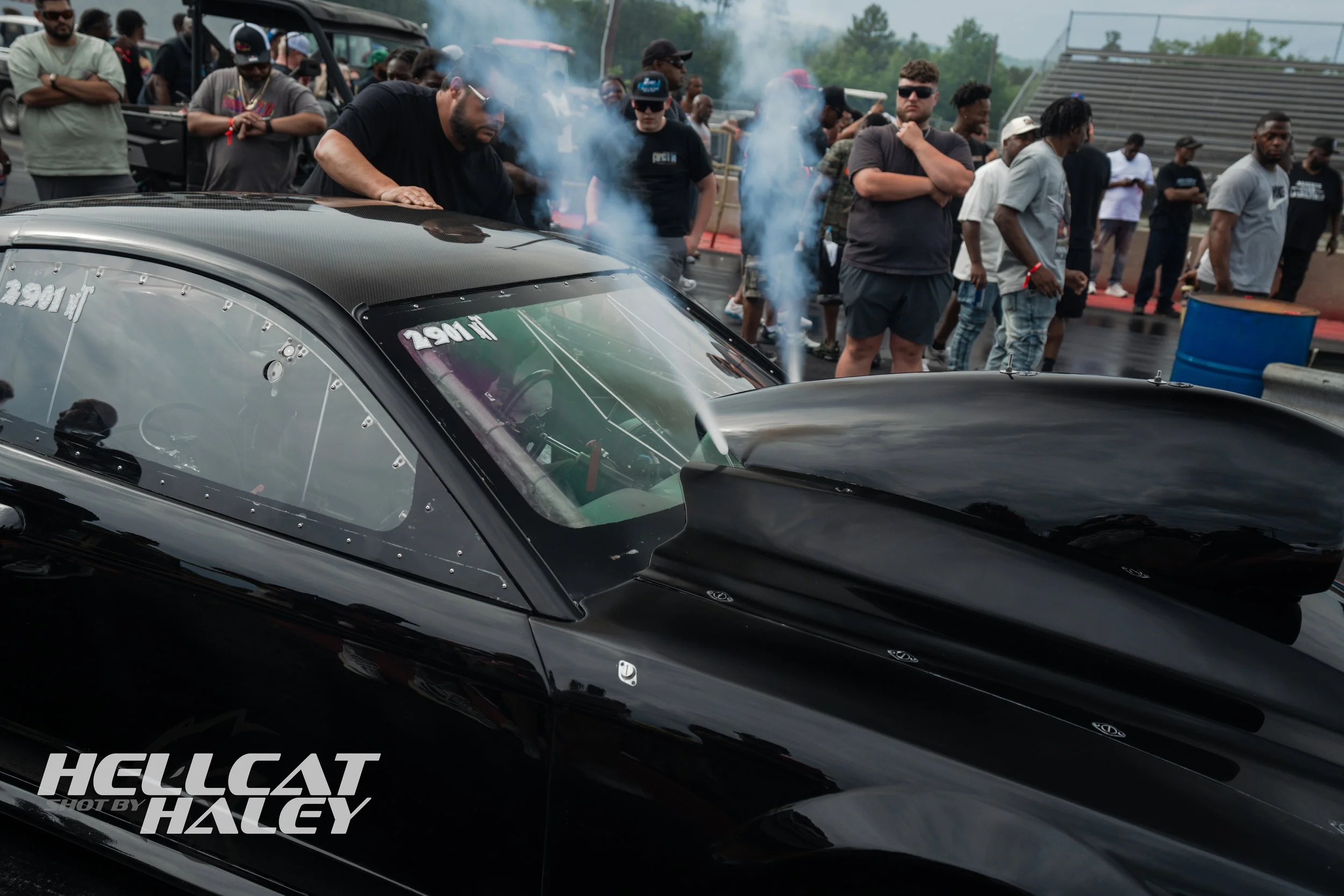 A group of people gathered around a black race car at a racing event, with some observing and others taking photos. The car has a reflective exterior, and a few spectators are visible in the background, standing near a track with a dirt surface and m