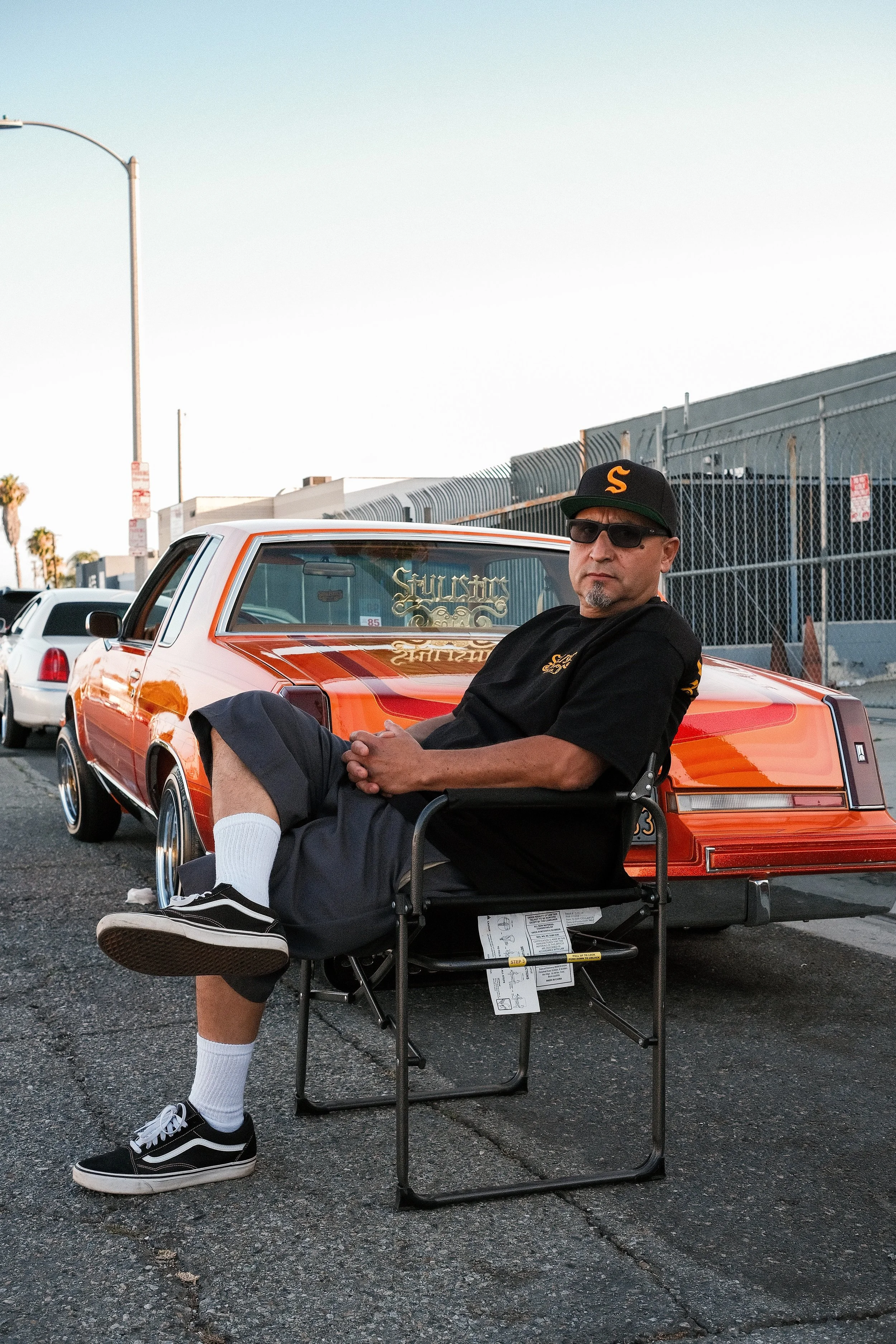 A man sitting in a wheelchair in a parking lot, wearing a black cap with an orange S, sunglasses, a black T-shirt, black shorts, and black sneakers with white soles. There is an orange and white vintage car behind him and a chain-link fence with indu