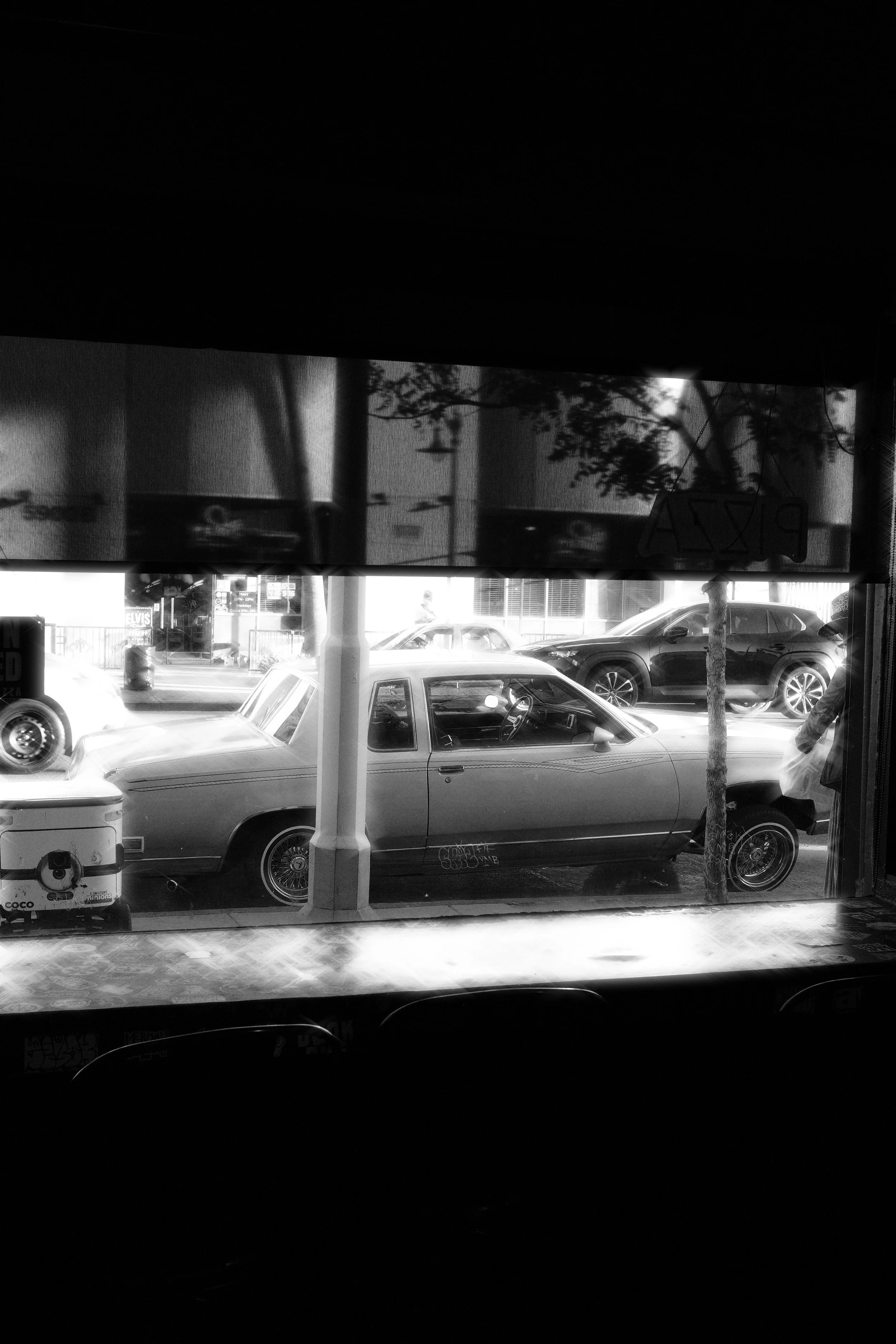 View through a window showing parked cars, a street scene, and a portion of a storefront with trees and street signs in the background.