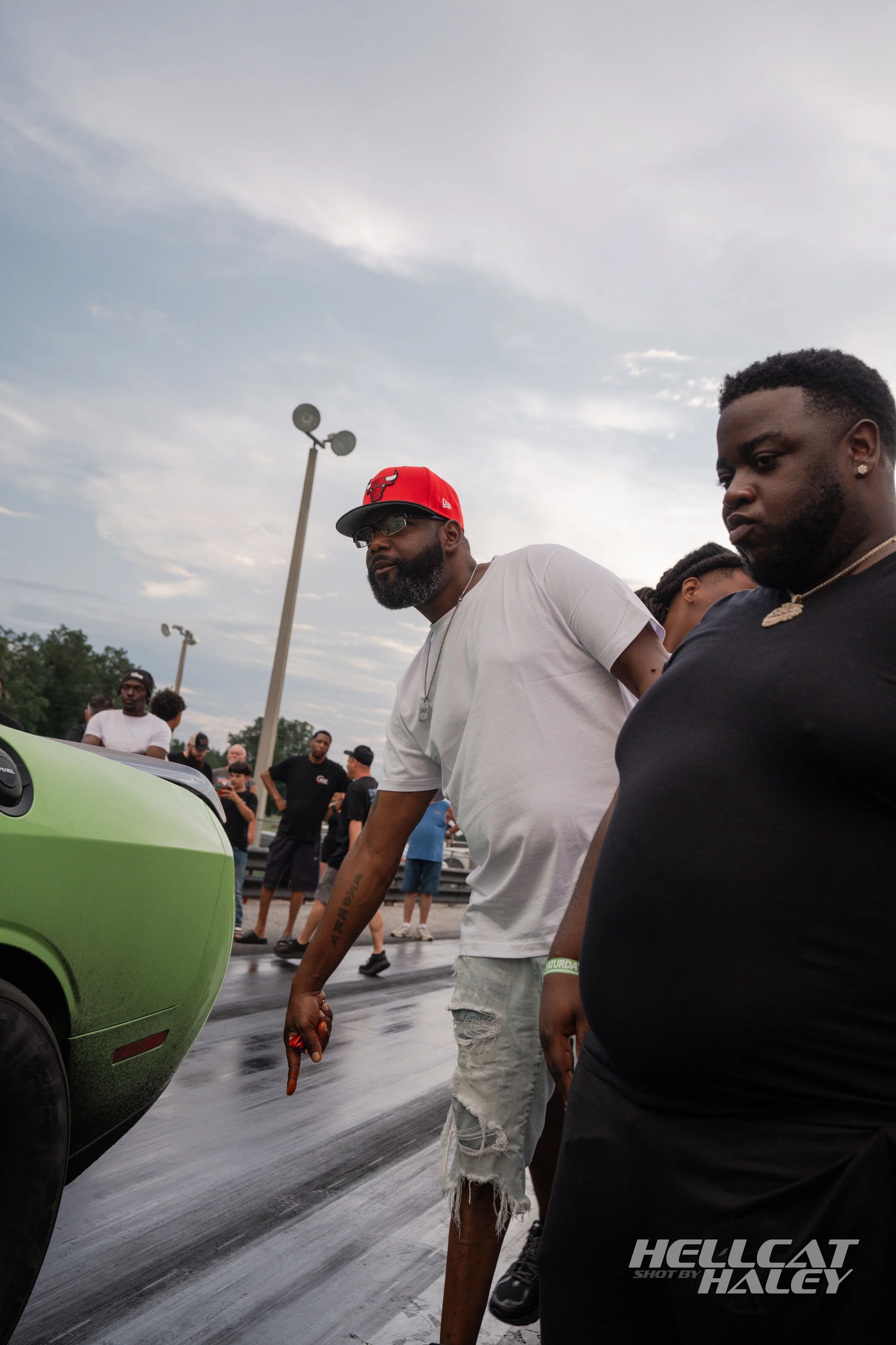 People gathered at an outdoor event, with two men in the foreground; one wearing a red cap and glasses, the other wearing a black shirt with a chain. They are standing near a green vehicle, with a cloudy sky and a crowd in the background.