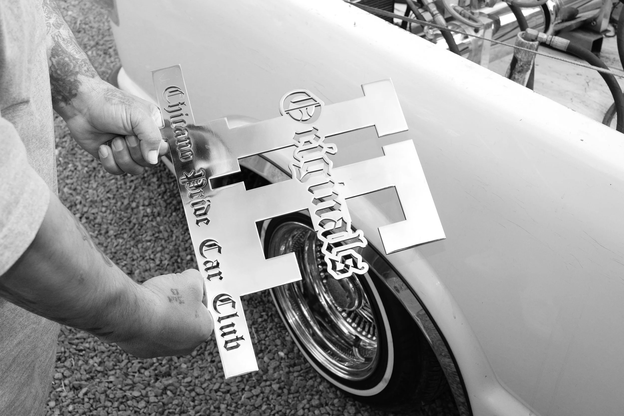 A person holding a large metallic cross-shaped sign with cut-out text, standing next to a vintage car with a chrome wheel.