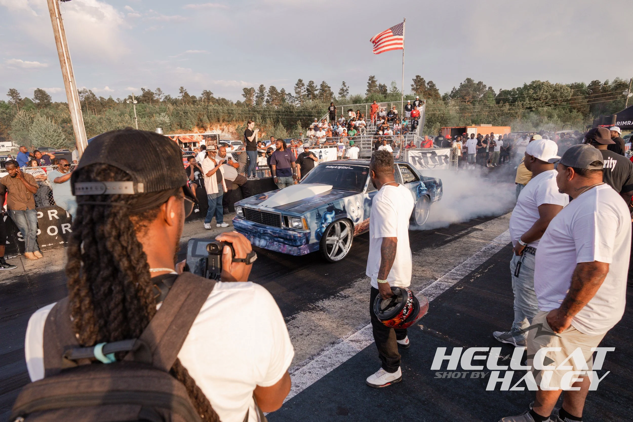 A blue race car performing a drift or burn out at a racing event, surrounded by a crowd of spectators and photographers. The event is outdoors with a partly cloudy sky and an American flag waving on a tall pole.