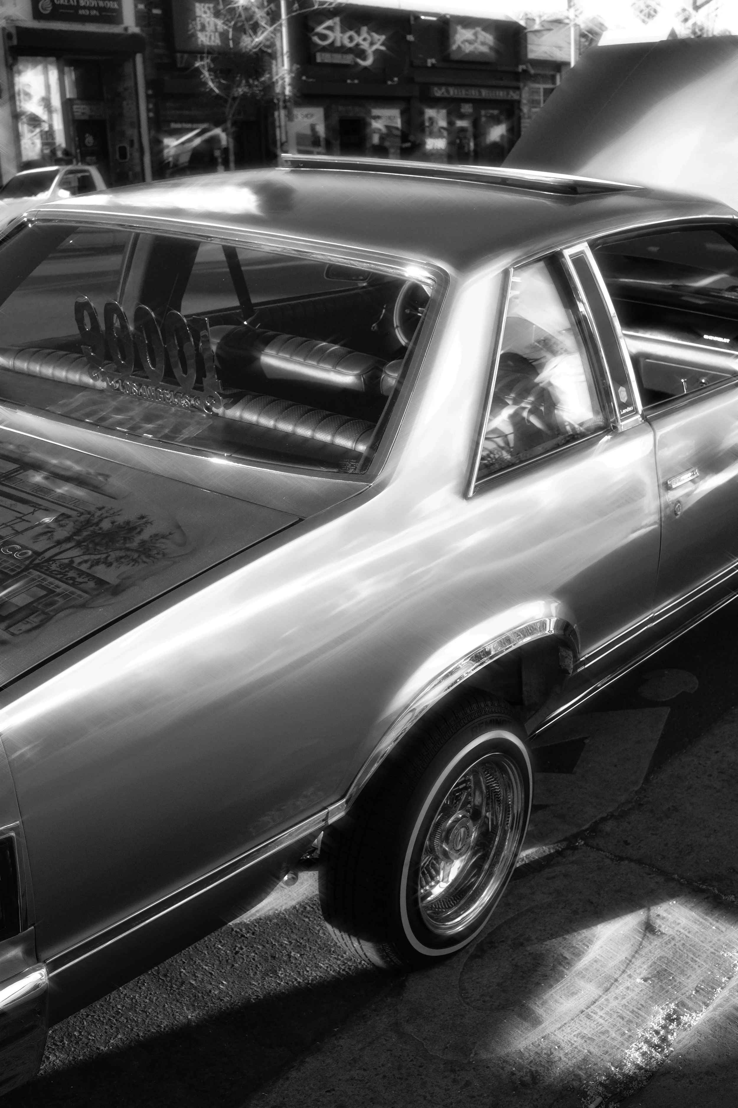 A black-and-white photo of a vintage car with a shiny exterior and a distinct rear end, parked on the street. The car's back window displays a decal with the numbers '500,' and the interior features a quilted upholstery. The background includes store