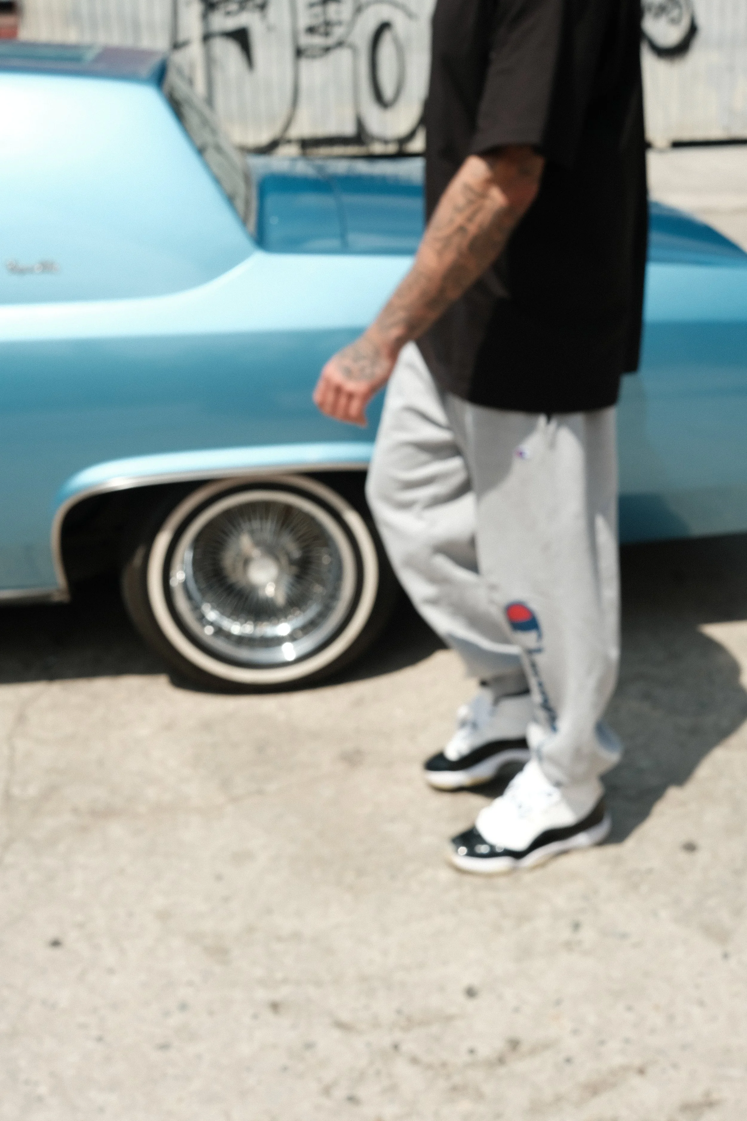 Person wearing a black shirt, white pants, and sneakers standing in front of a vintage blue car.