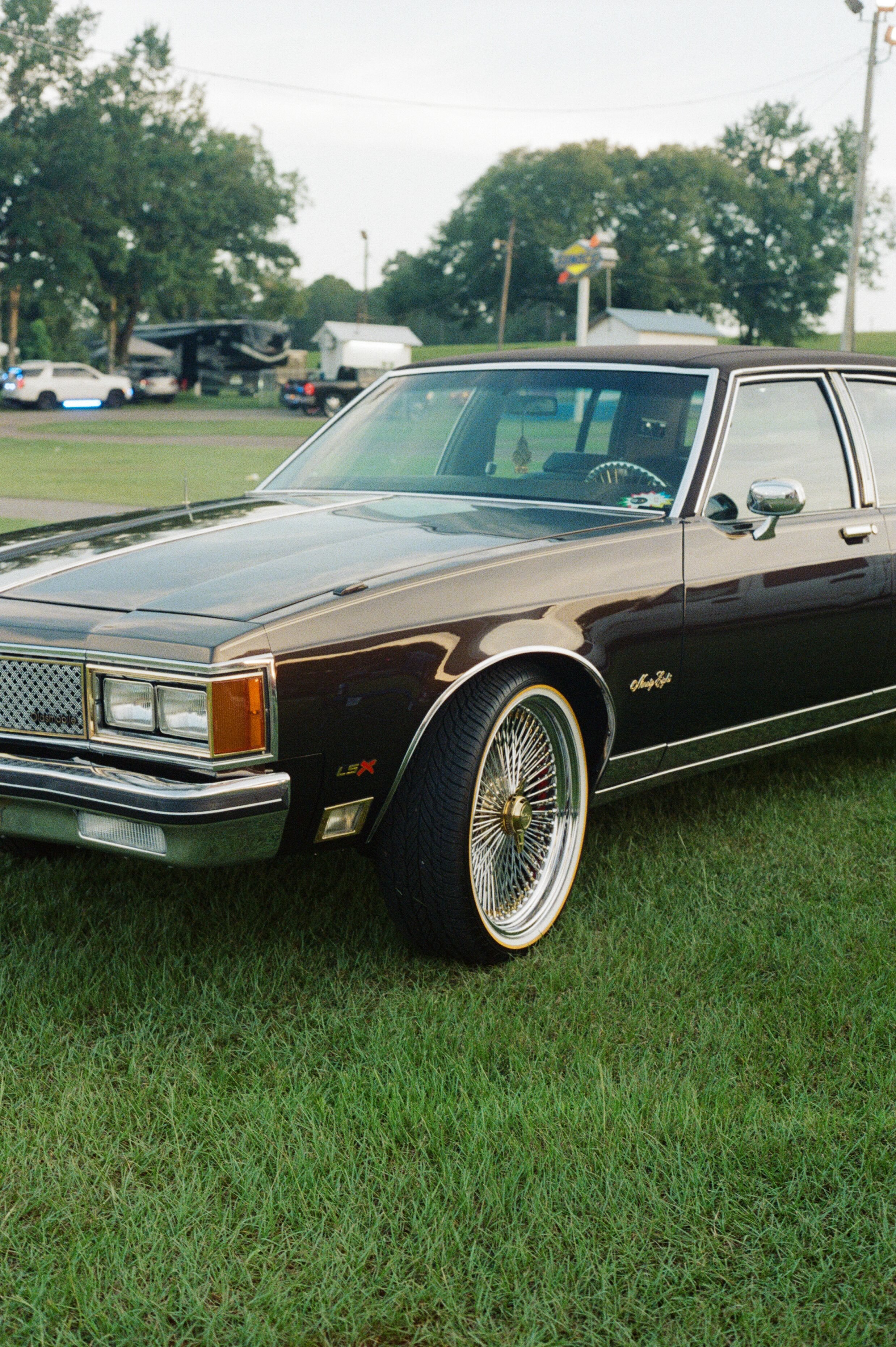 A vintage black Cadillac Eldorado parked on a grassy field with other vehicles and trees in the background, under a cloudy sky.