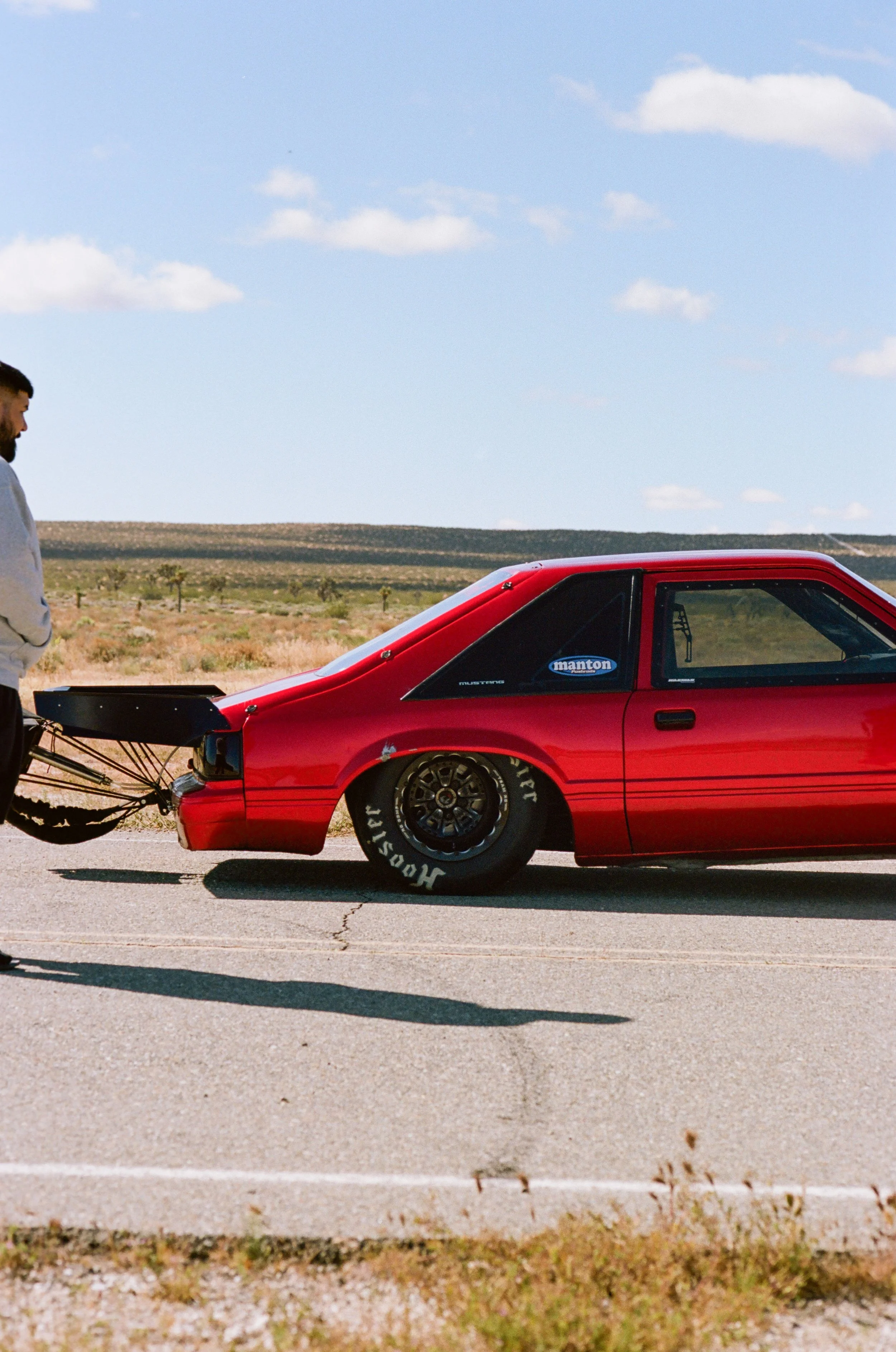 A man in a light gray hoodie stands beside a red race car with a black spoiler, in a desert landscape with sparse vegetation and a blue sky with scattered clouds.