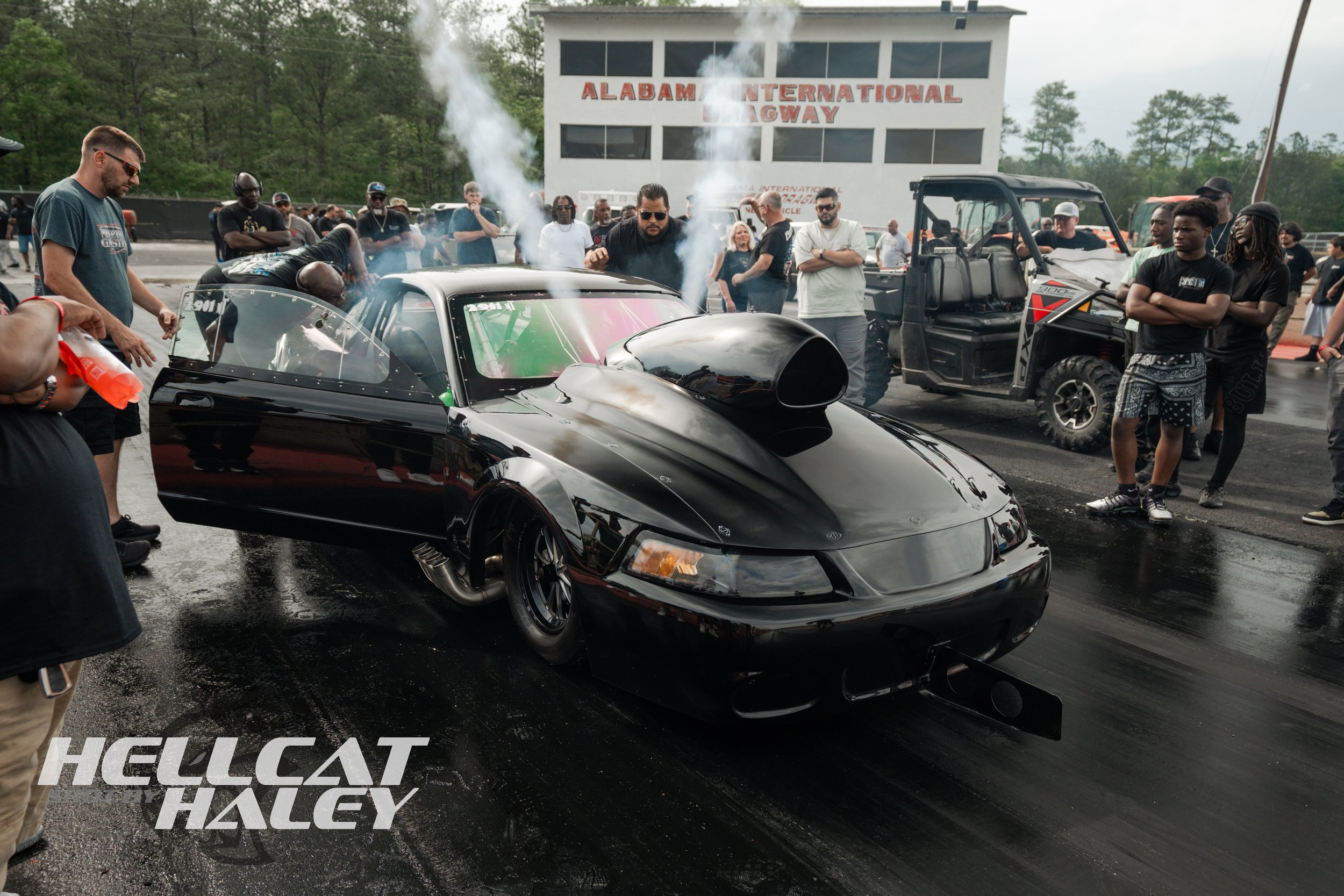 A black race car with a large hood scoop and tinted windows is on a drag strip, surrounded by a crowd of people, some taking photos and others watching. There is smoke coming from the car's rear tires. In the background, a white building with red let