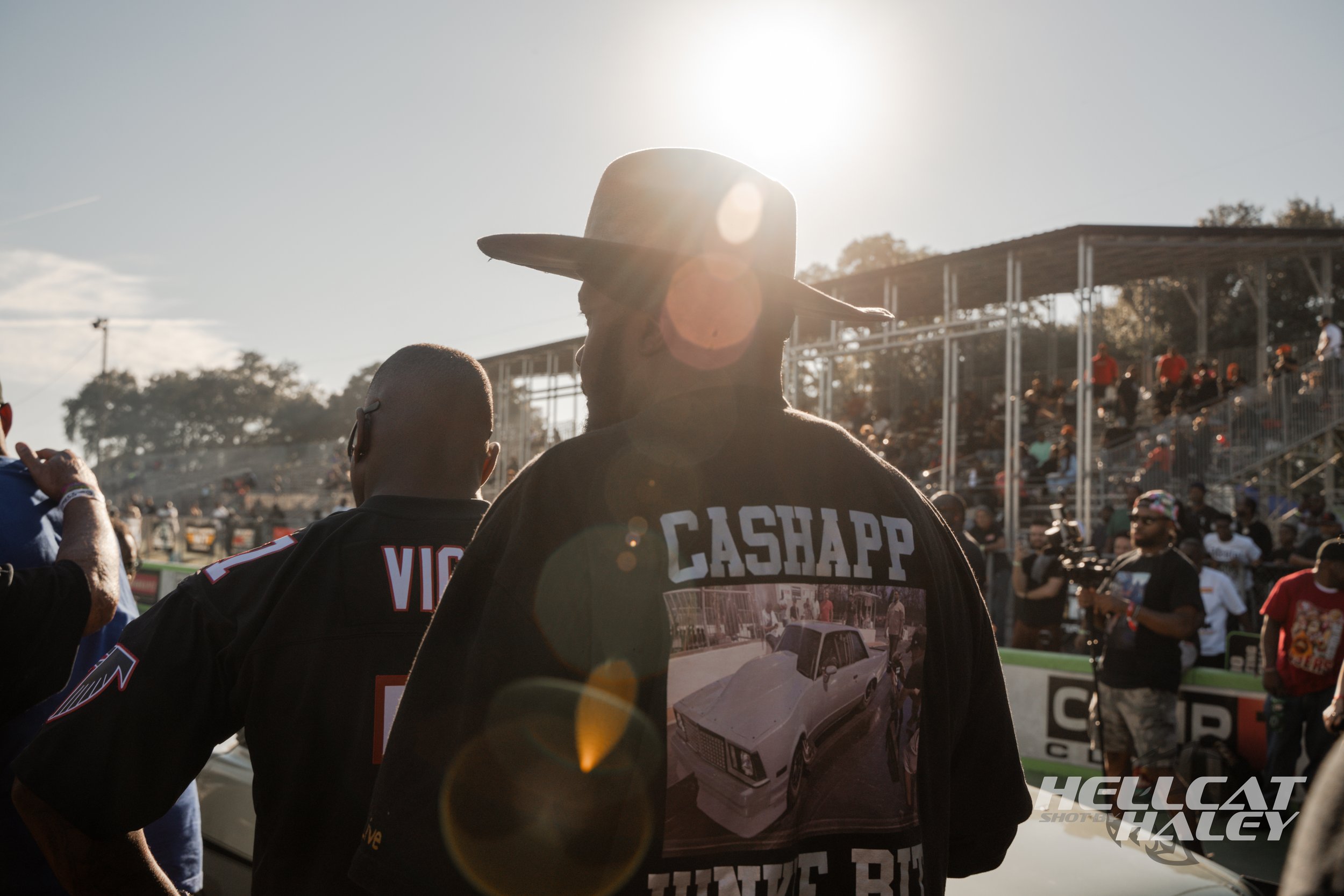 Two men at a racing event, with one wearing a cowboy hat and the other with a t-shirt featuring a car image, crowd in the background under bright sunlight.
