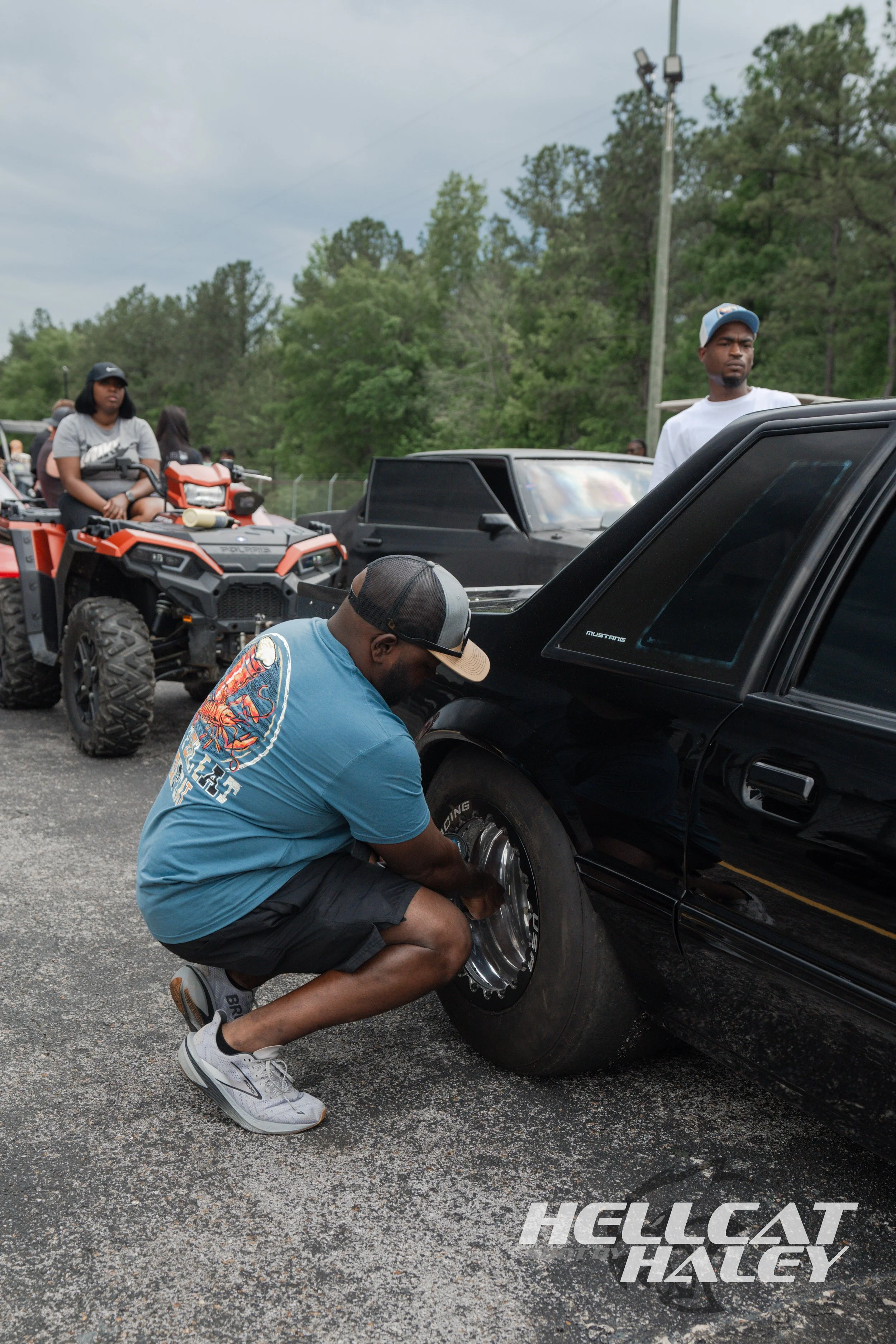 A man crouching down changing a black tire on a black car, with several people and vehicles, including a quad bike, in the background outdoors.