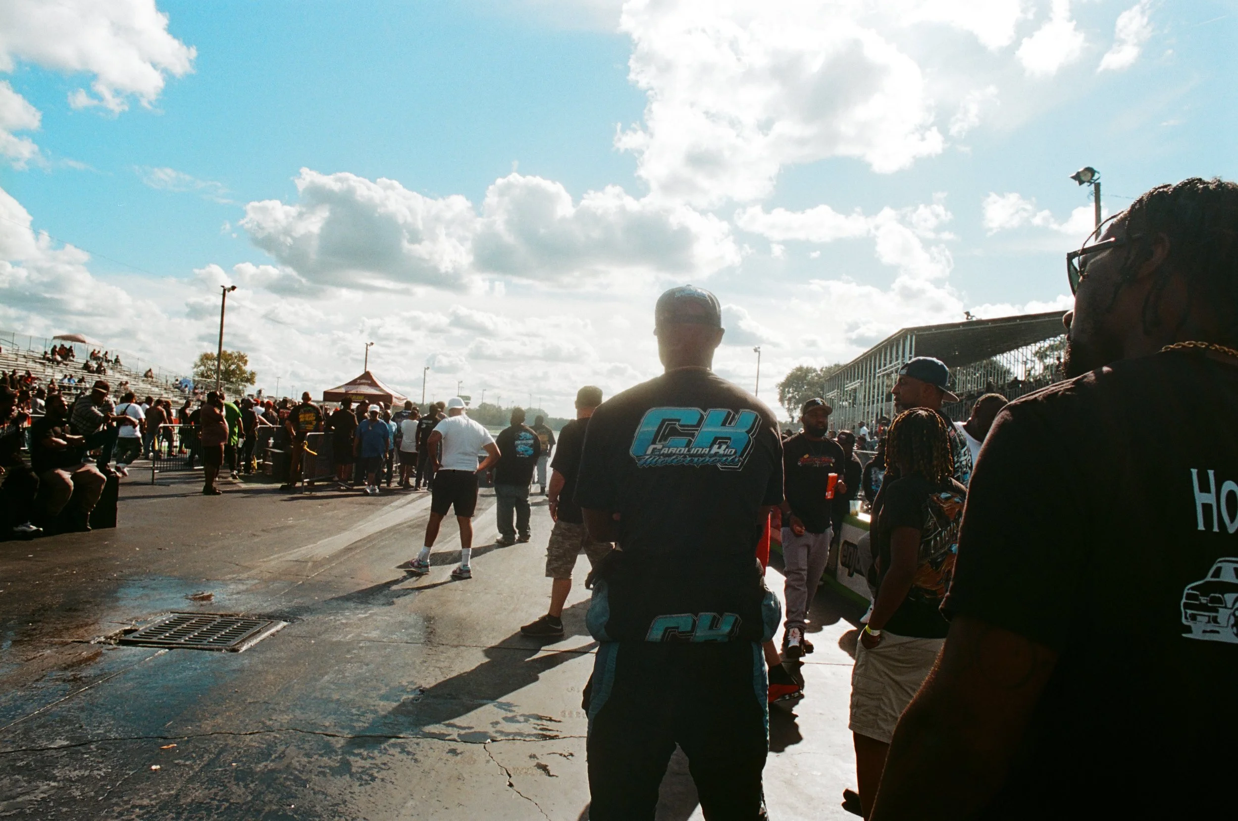 Crowd gathering at an outdoor event on a sunny day with a partly cloudy sky, bleachers on the left, and people standing near a race track.