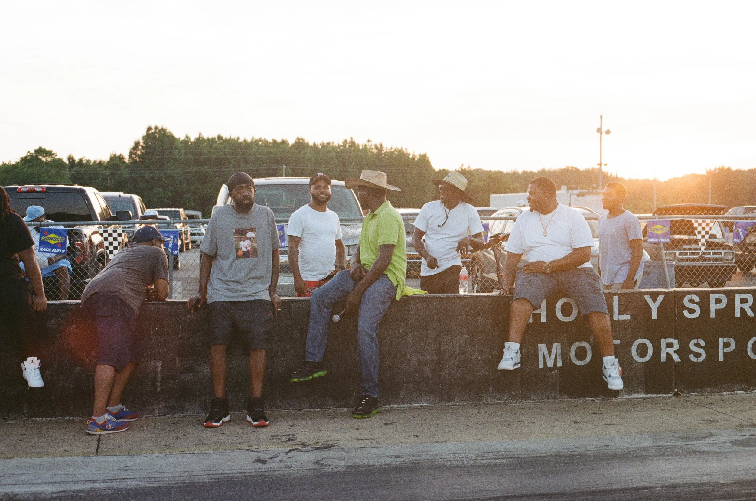 Group of six men standing and sitting near a racetrack fence during sunset, with cars and racing banners in the background.