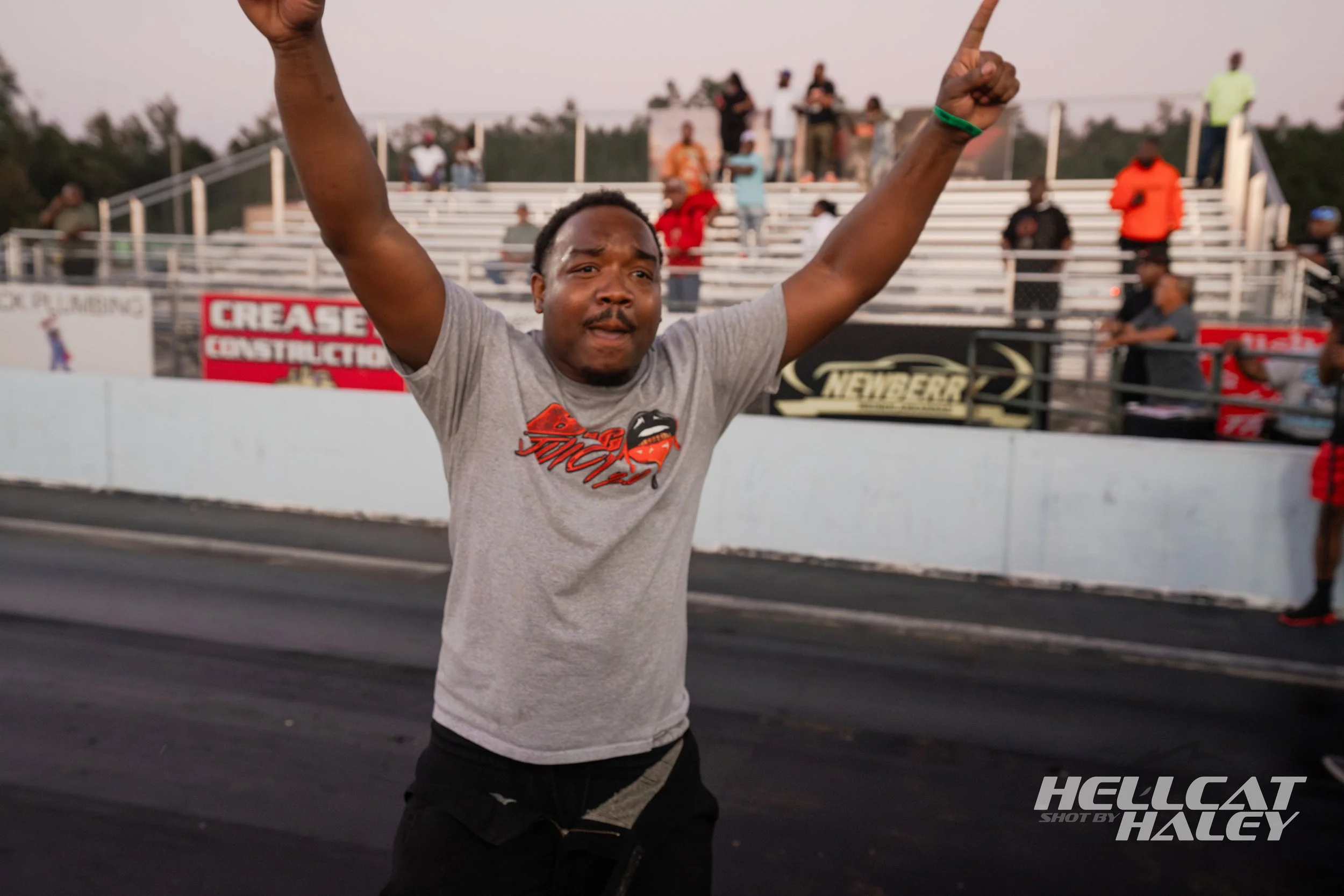 Man celebrating at a race track with arms raised and fists closed, crowd in the background.