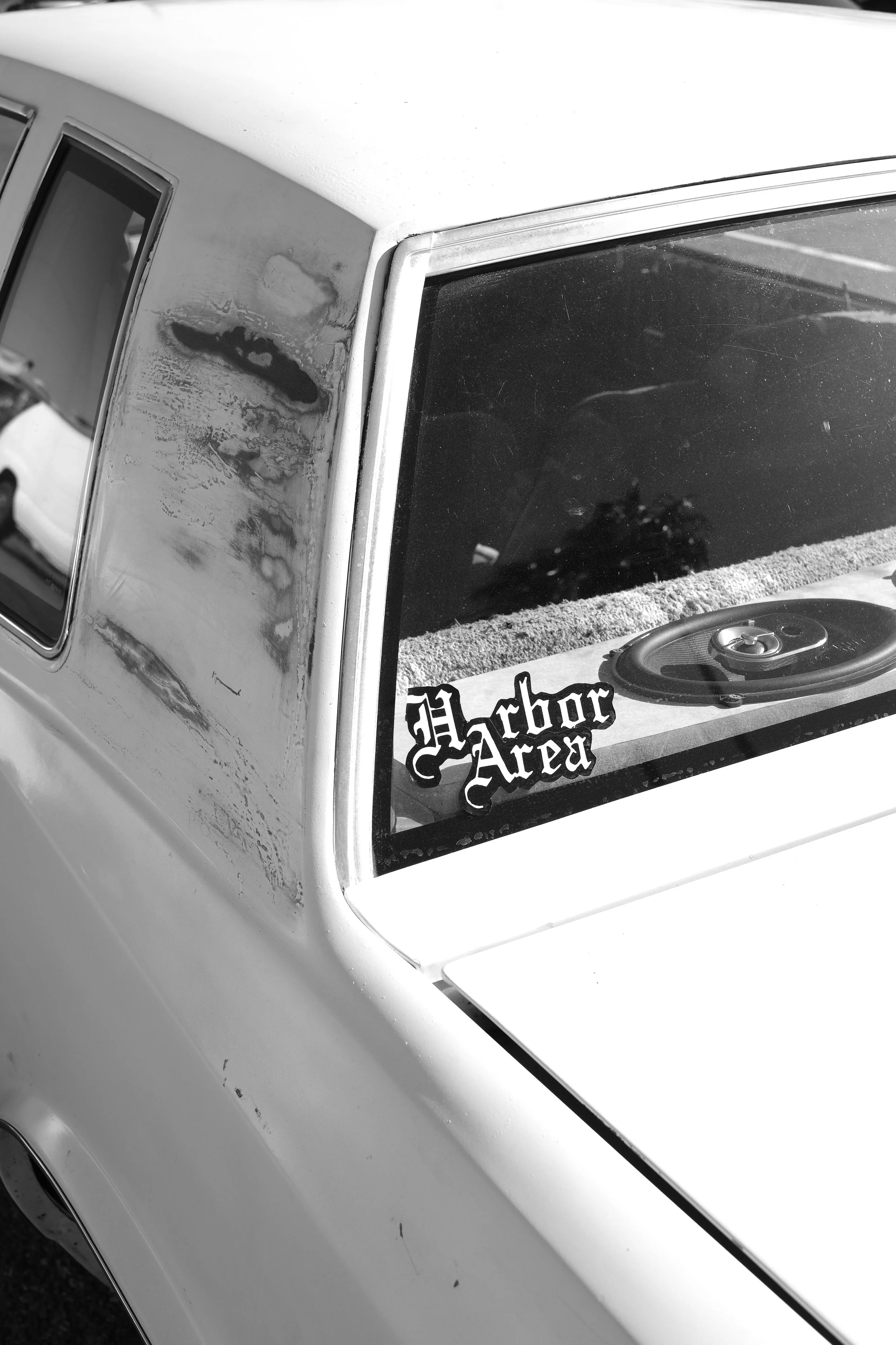 Close-up of the front windshield and side window of a vintage car with a sticker reading 'Harbor Area' near the dashboard, some dirt and scratches on the window, and a speaker on the dashboard.