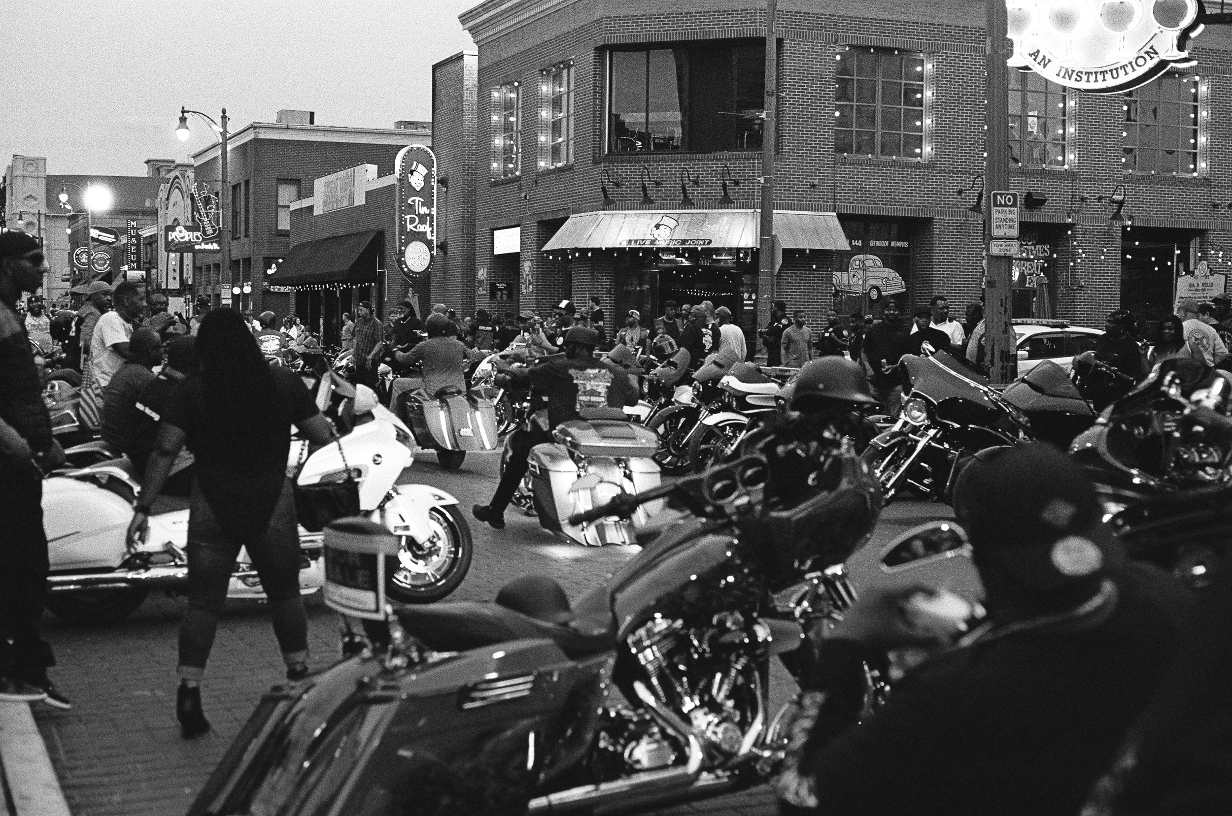 A black-and-white photo of a crowded street scene with numerous parked motorcycles and people walking around or riding them. In the background, there are brick buildings with illuminated signs and string lights, indicating an urban nightlife setting.