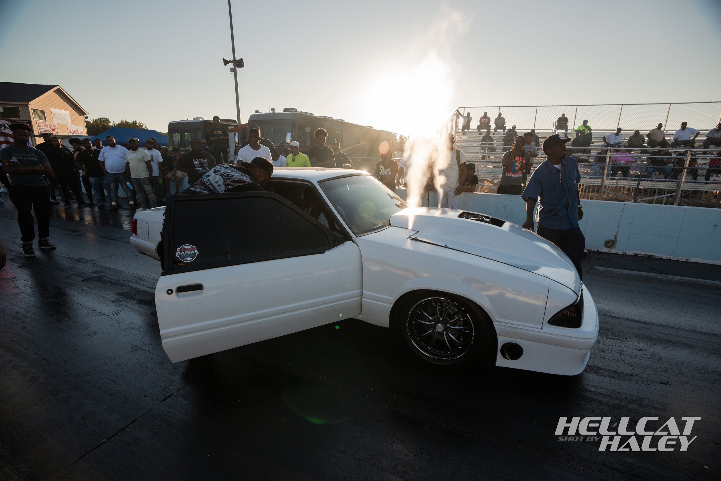 A drag racing event at a racetrack with a white car at the starting line and a crowd of spectators in the background.