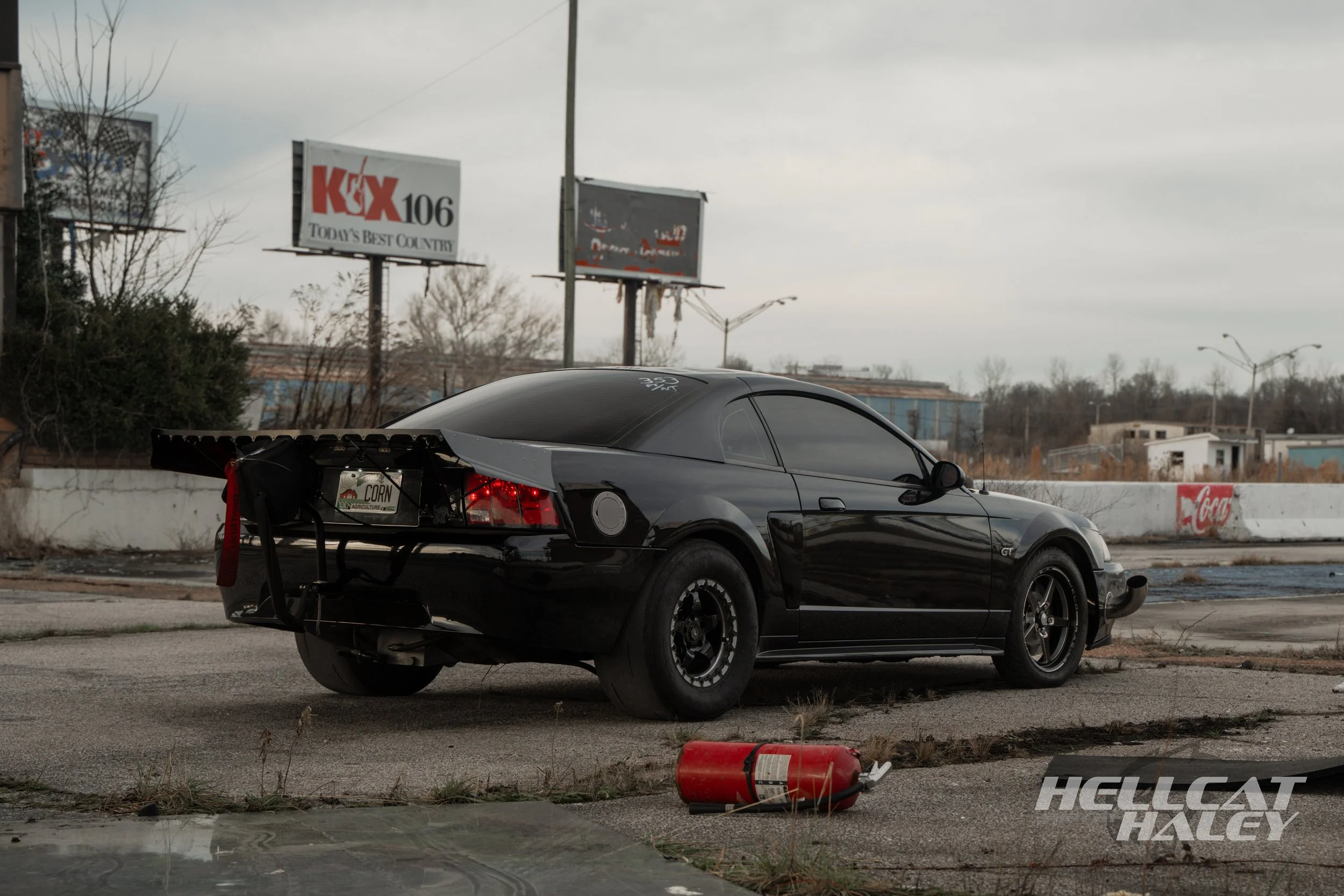 A black sports car with a rear wing, parked on the side of the road, with a damaged rear bumper. An overturned fire extinguisher and debris are on the ground nearby. In the background, there are billboards, one advertising KX 106 radio station and an