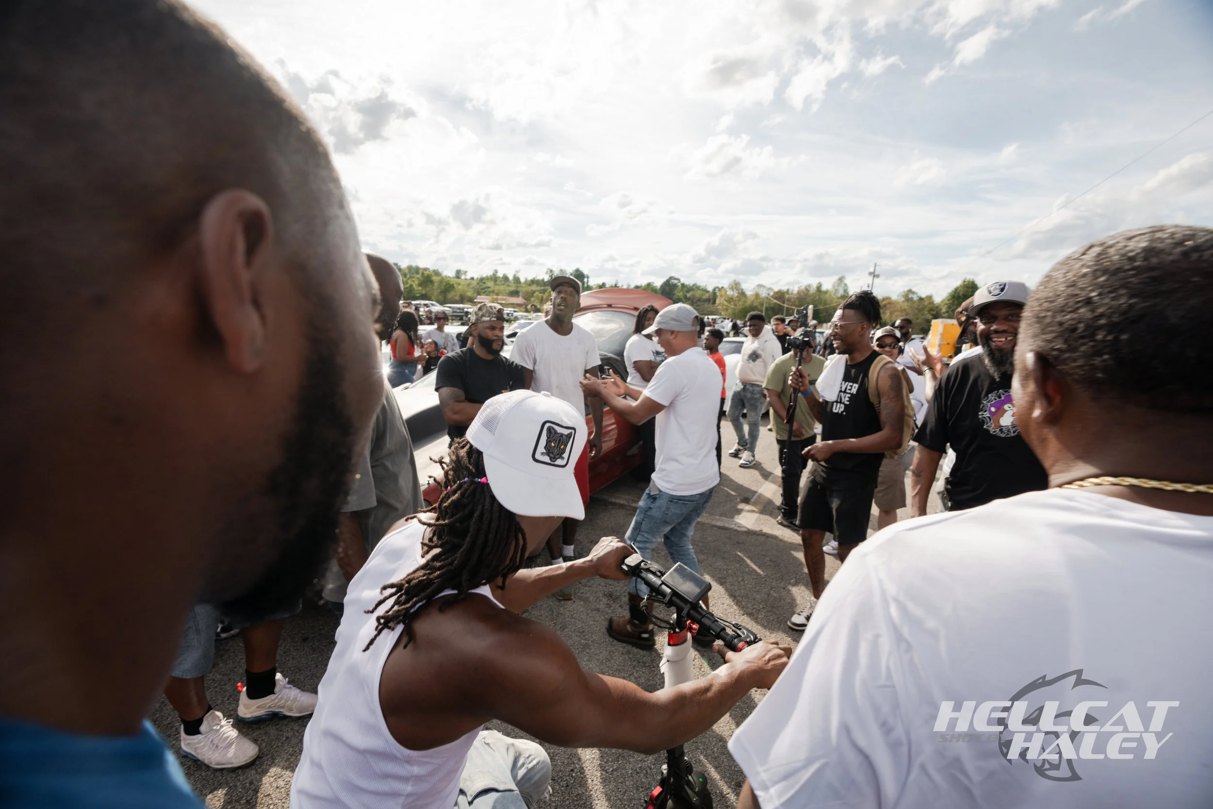 A group of people gathered outdoors in a parking lot, some engaging in conversation, with cars visible in the background and cloudy sky overhead.
